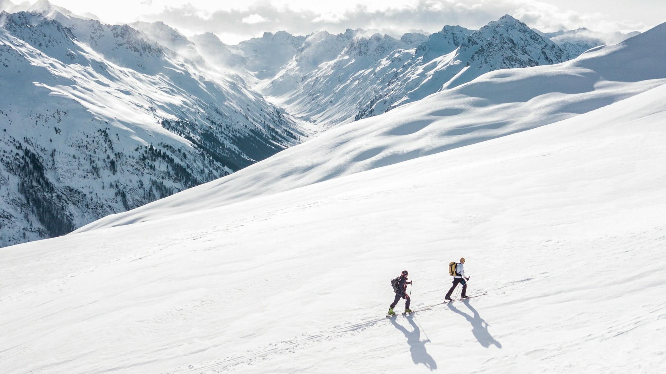 Two skiers walking up a very snowy hill on skis. There are many snowy mountains in the background.