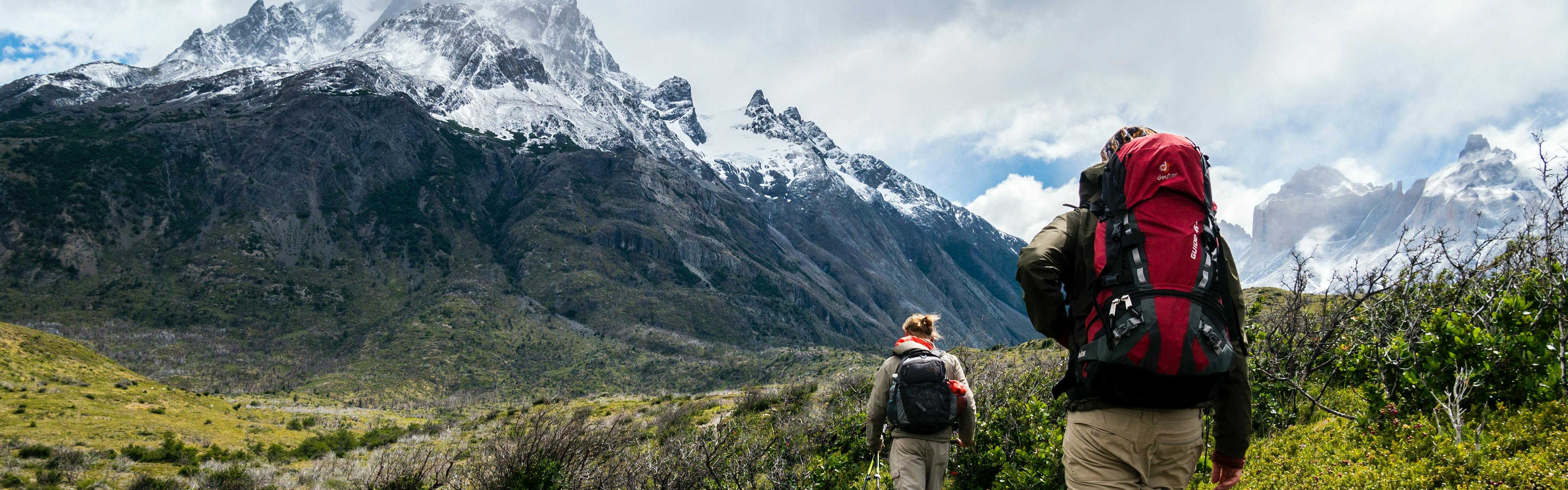 Two backpackers walk away from the camera towards snow-covered peaks. 