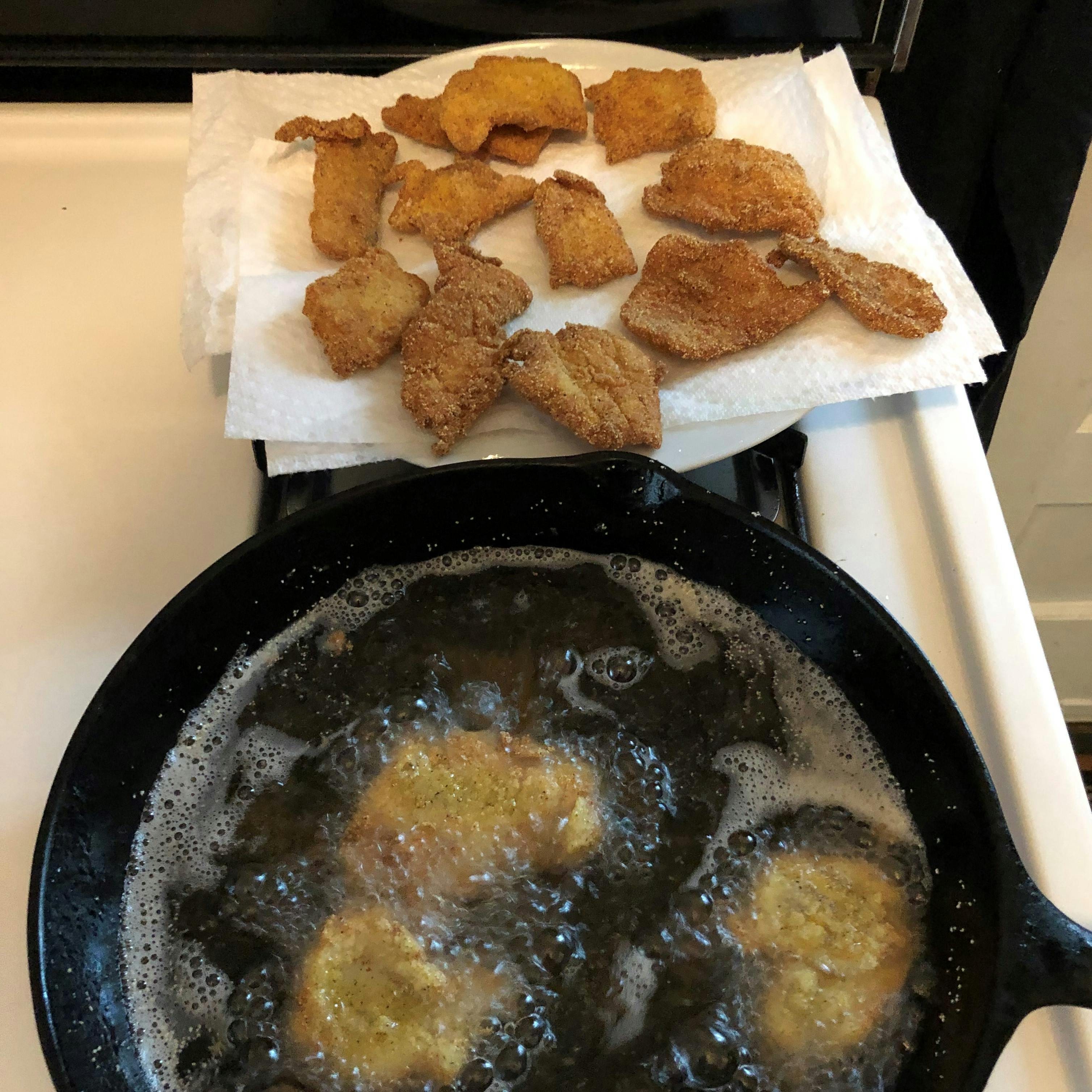 Fried crappie sits on a plate and some is frying in a cast-iron pan. 