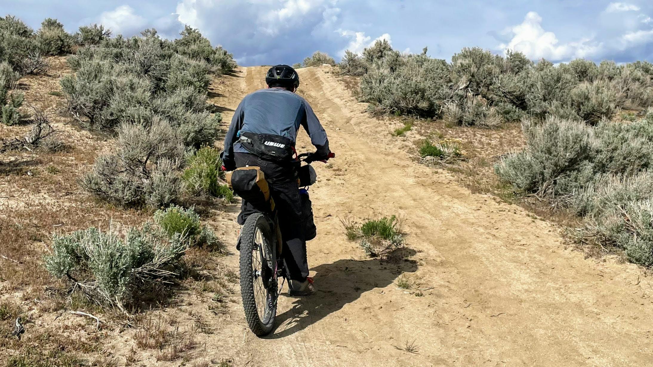 A biker rides up a dirt trail. There is sagebrush on the sides of the trail.