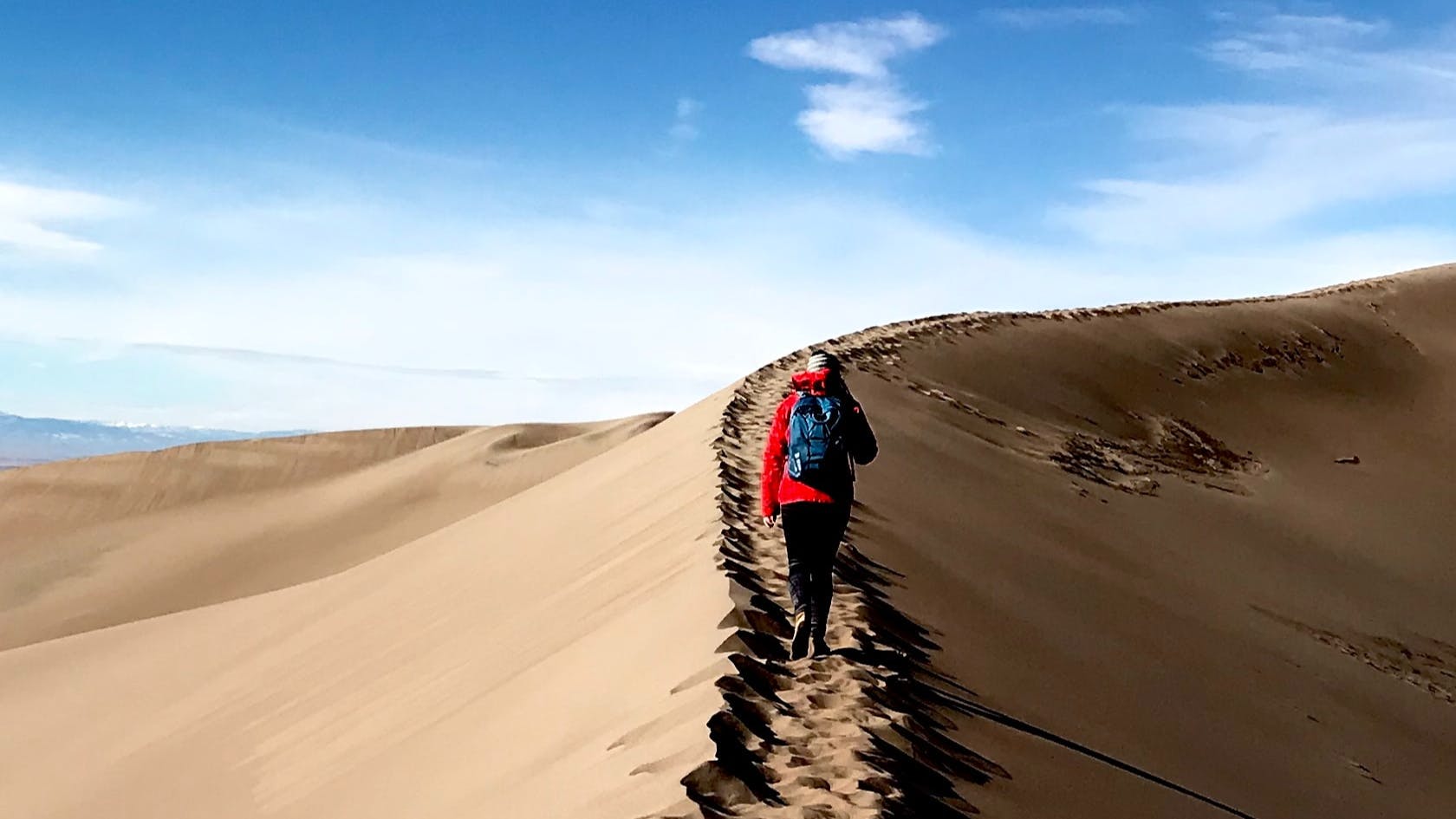 Someone in a red jacket walks on the ridge of a sand dune with their back towards the camera. The sky is bright blue ahead of them. 