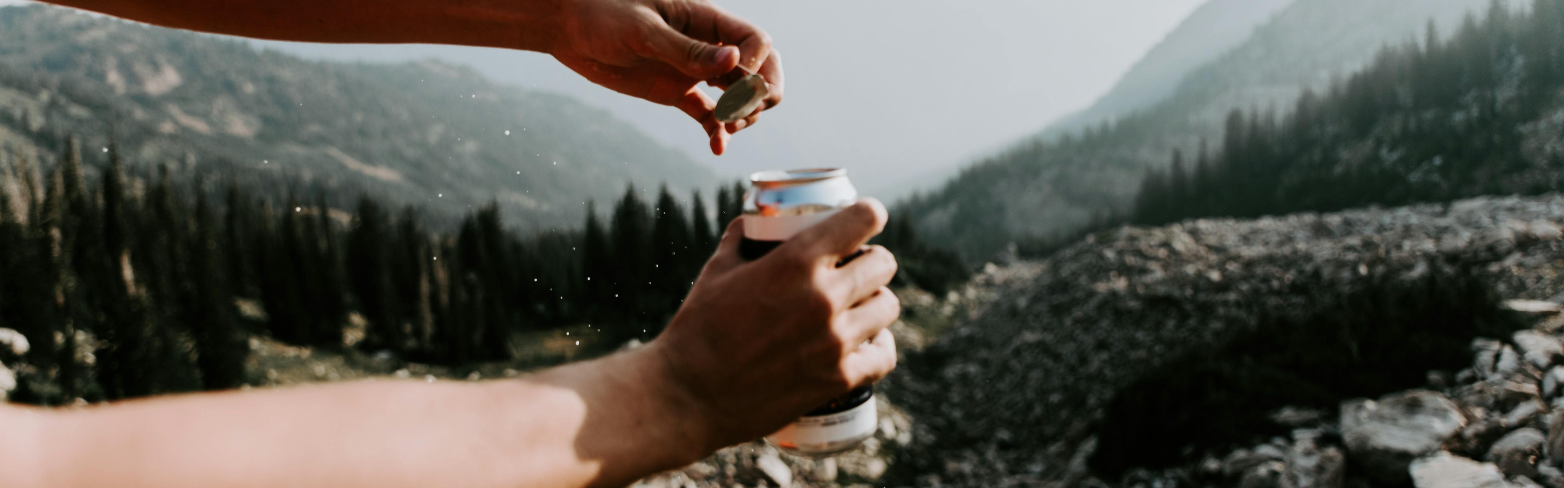 A hand pulling the top off of a can. There are mountains in the background. 