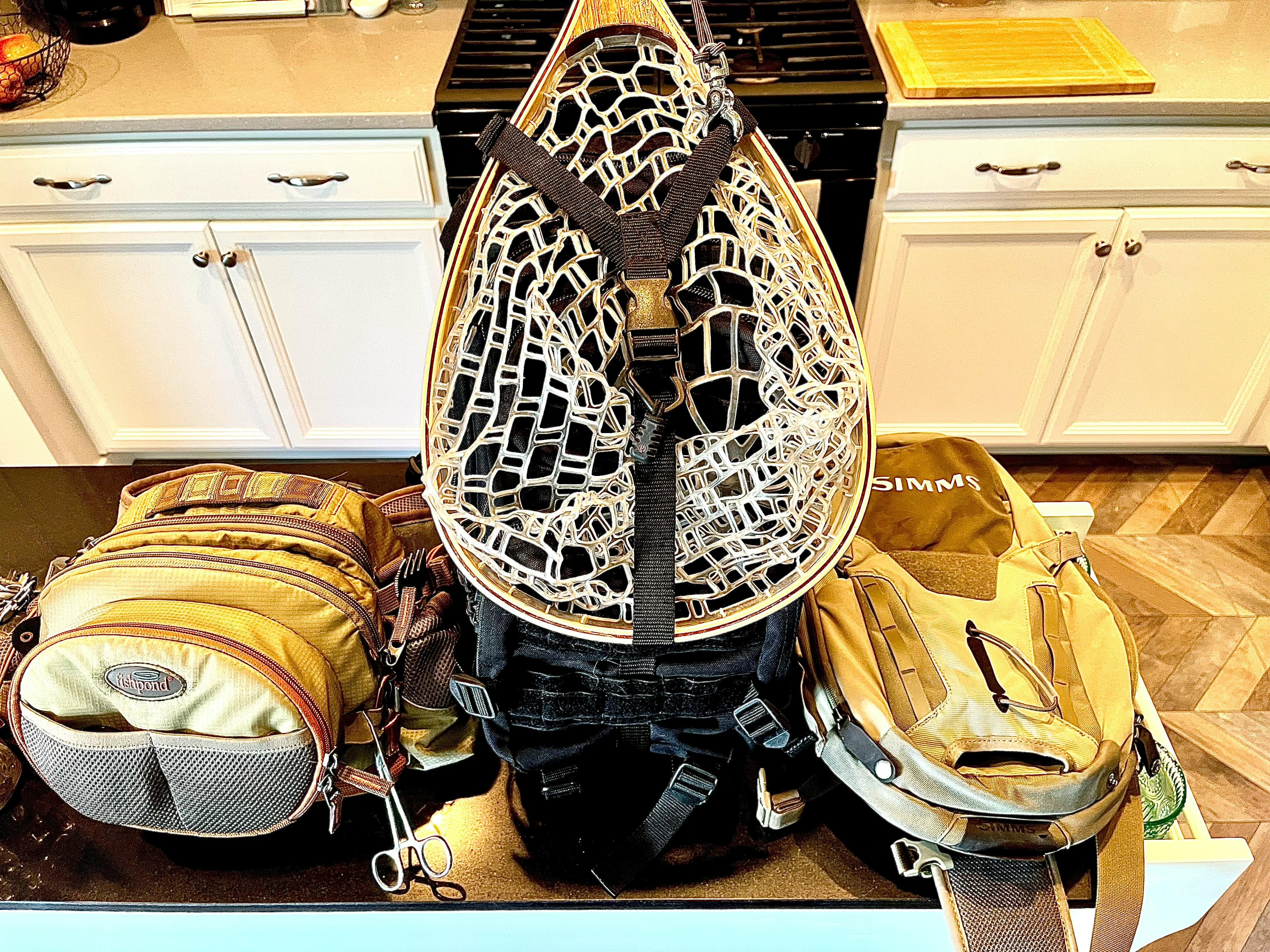 A fly fishing pack is laying in a kitchen with all the gear needed for fly fishing, such as a net, attached to the outside. 