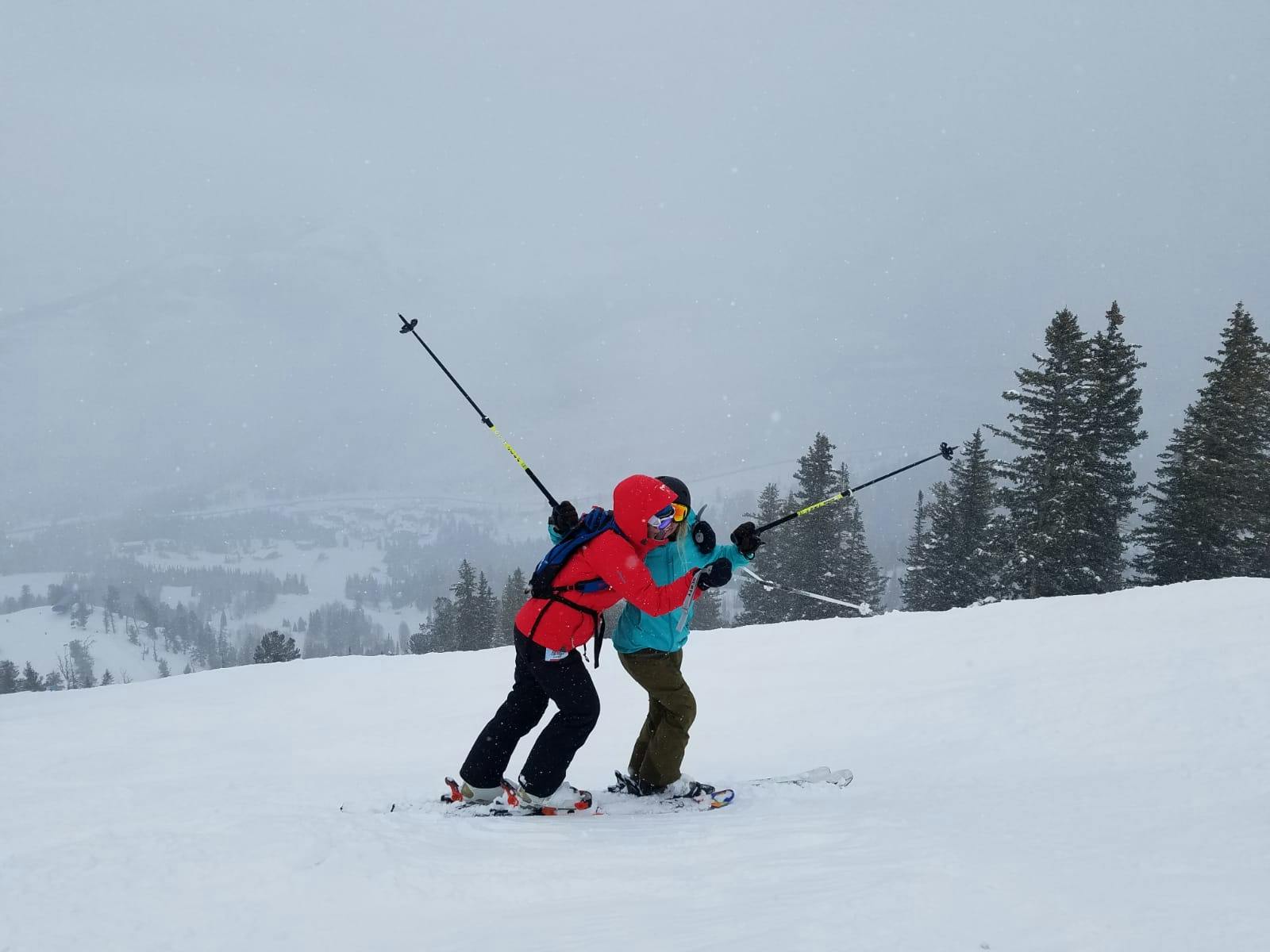 Two skiers on a hill hug each other with their poles in the air.