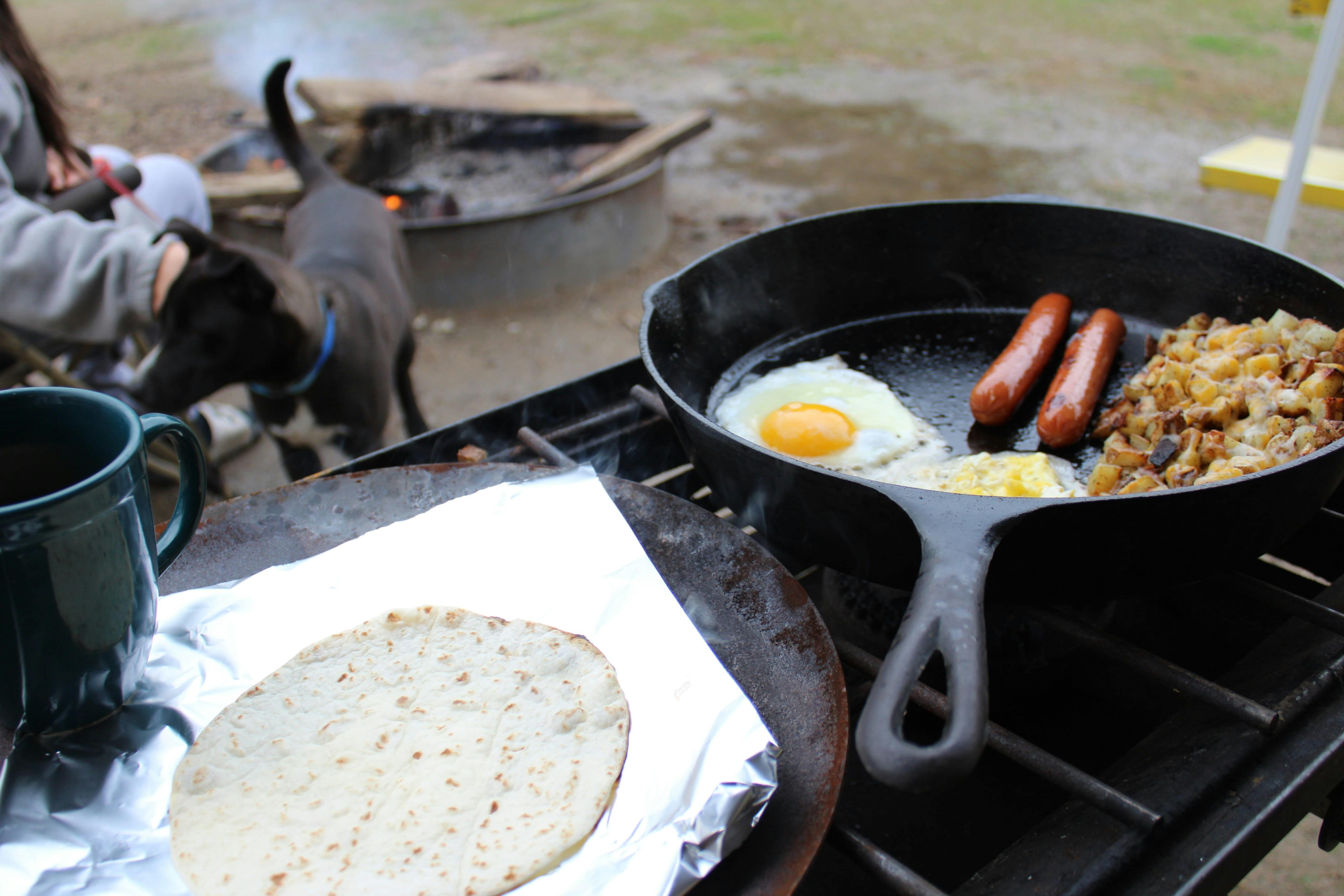 A cast-iron pan with fried eggs, sausages, and a scramble next to tin foil with a tortilla on it, all on a grill. 