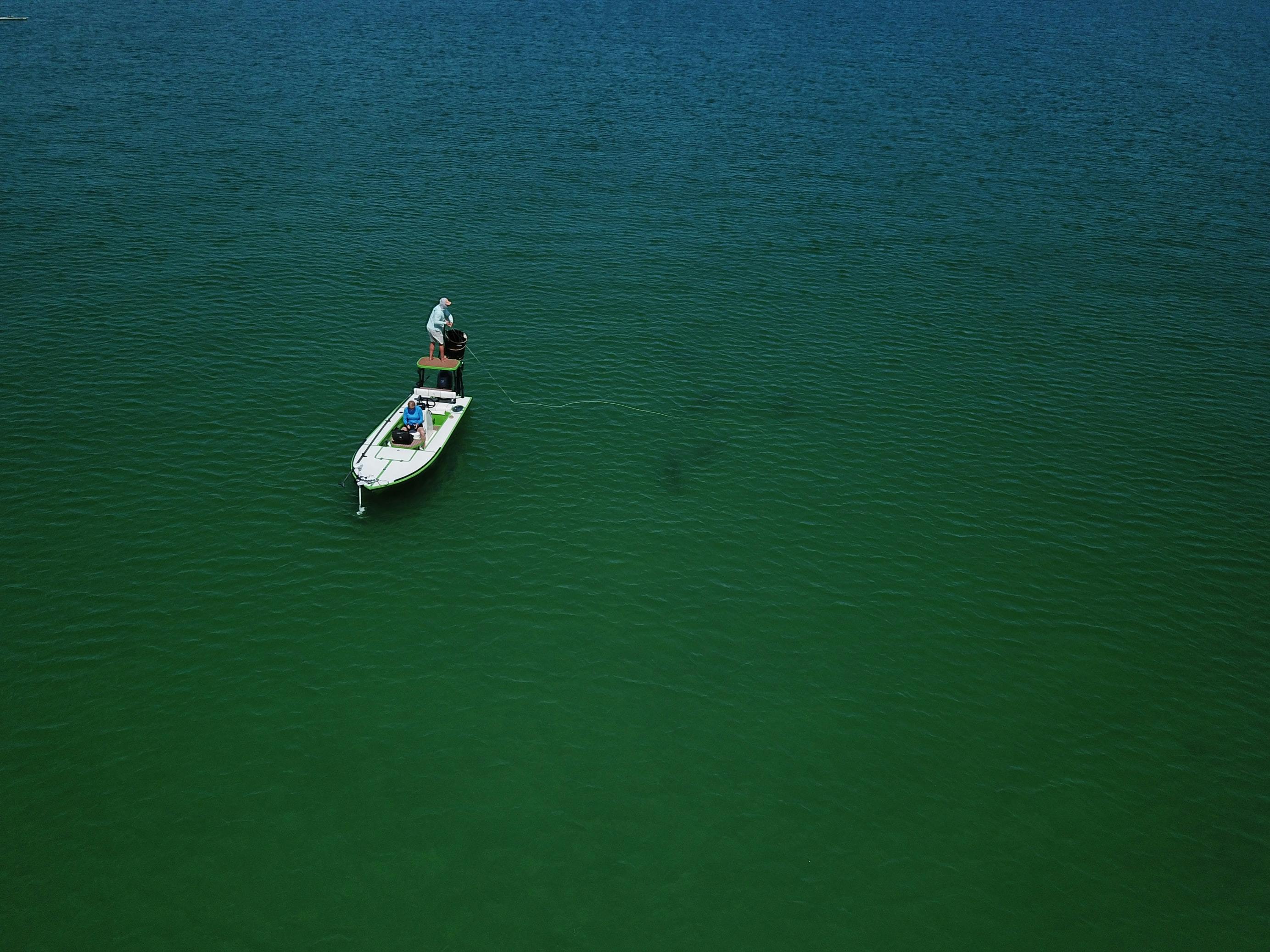 An aerial shot of someone standing on a platform on a boat above shallow water. They are casting with their fly rod.