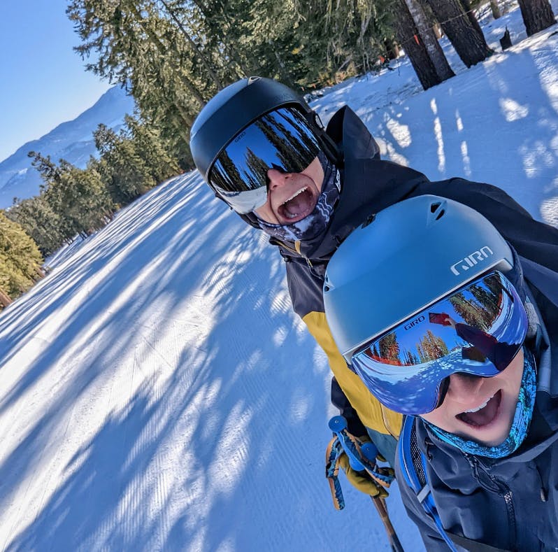 A selfie of two people on a snowy ski trail. They are both wearing helmets and goggles and smiling at the camera. 