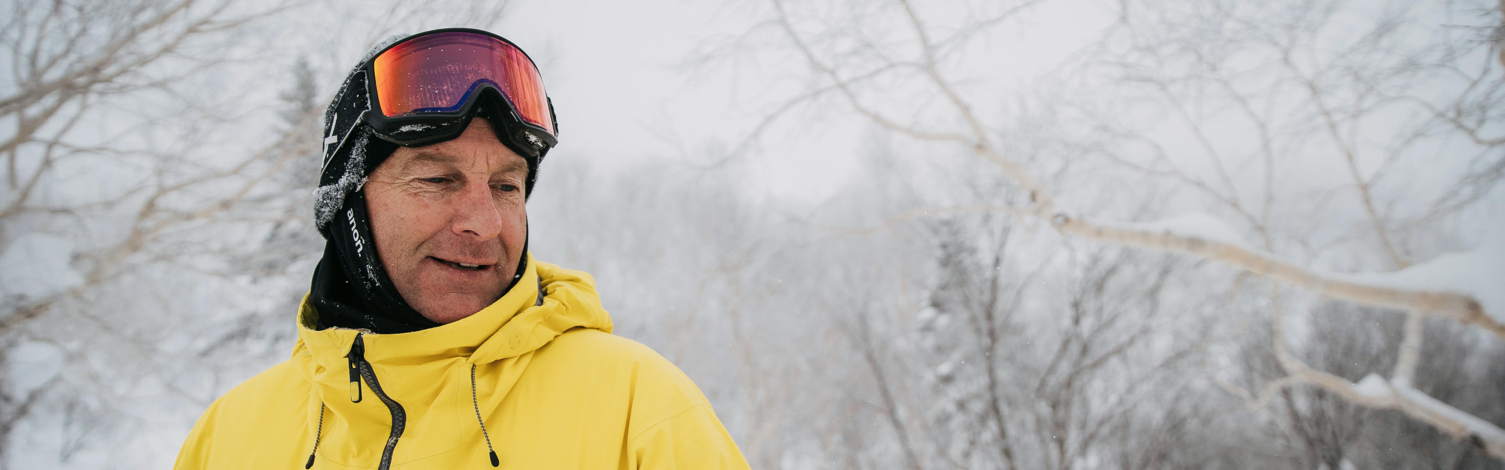 Dave Downing smiles and looks down while wearing a yellow jacket and googles. The landscape behind him is snowy and gray.