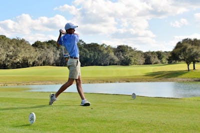Golfer in blue shirt and shorts holds finish of golf swing with golf course in background