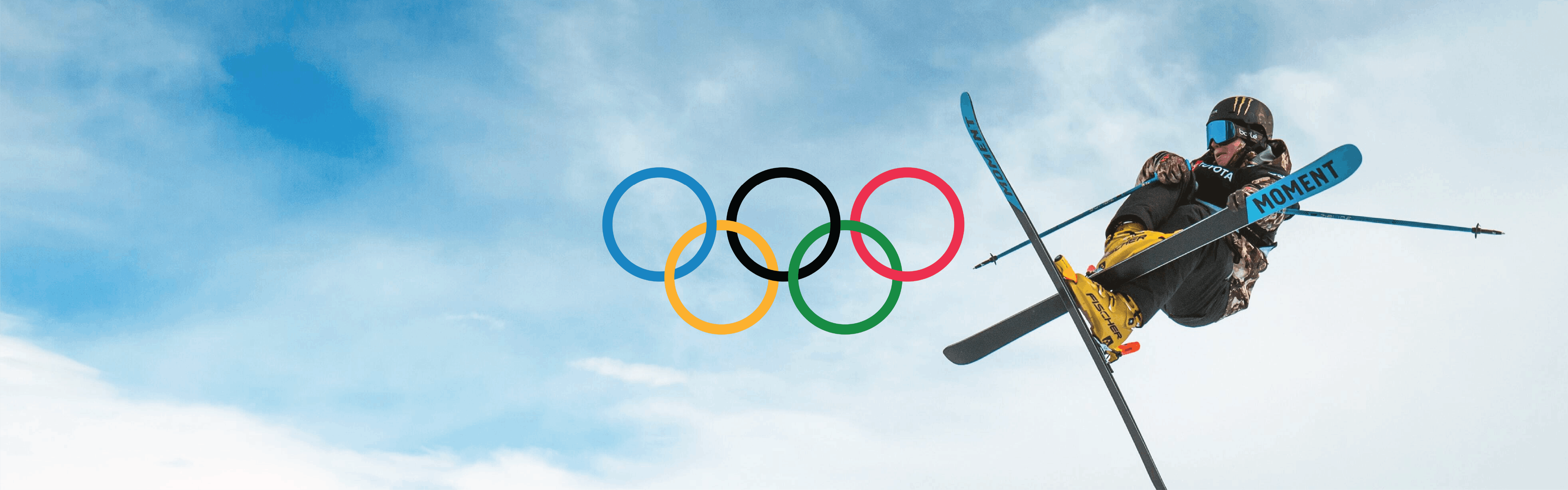 A skier jumps in the air and crosses his skis. The sky behind him is blue and dusted with clouds. The Olympic rings are added on top.