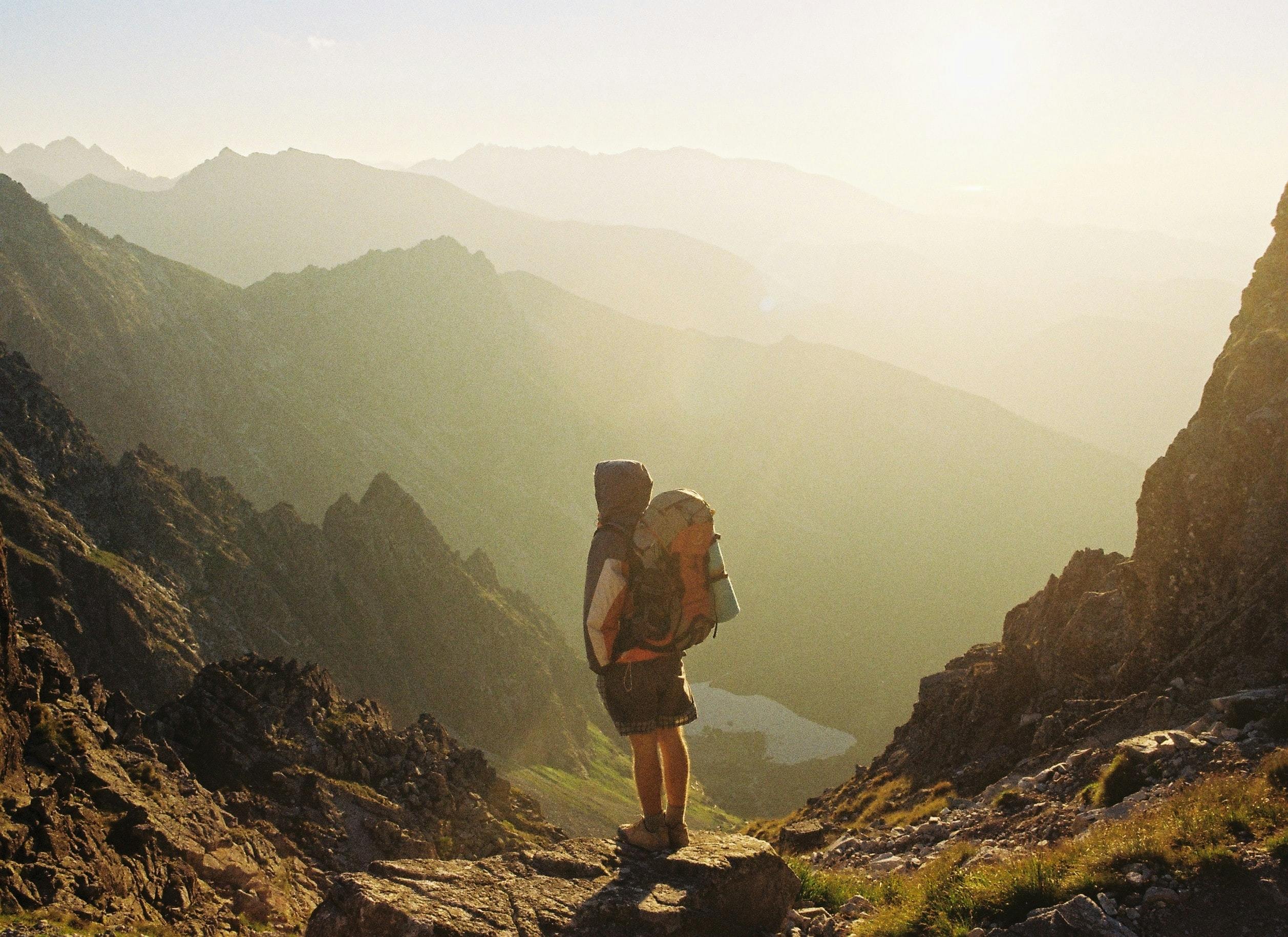 A backpacker stands on a ridge overlooking a lake below and jagged peaks ahead. 