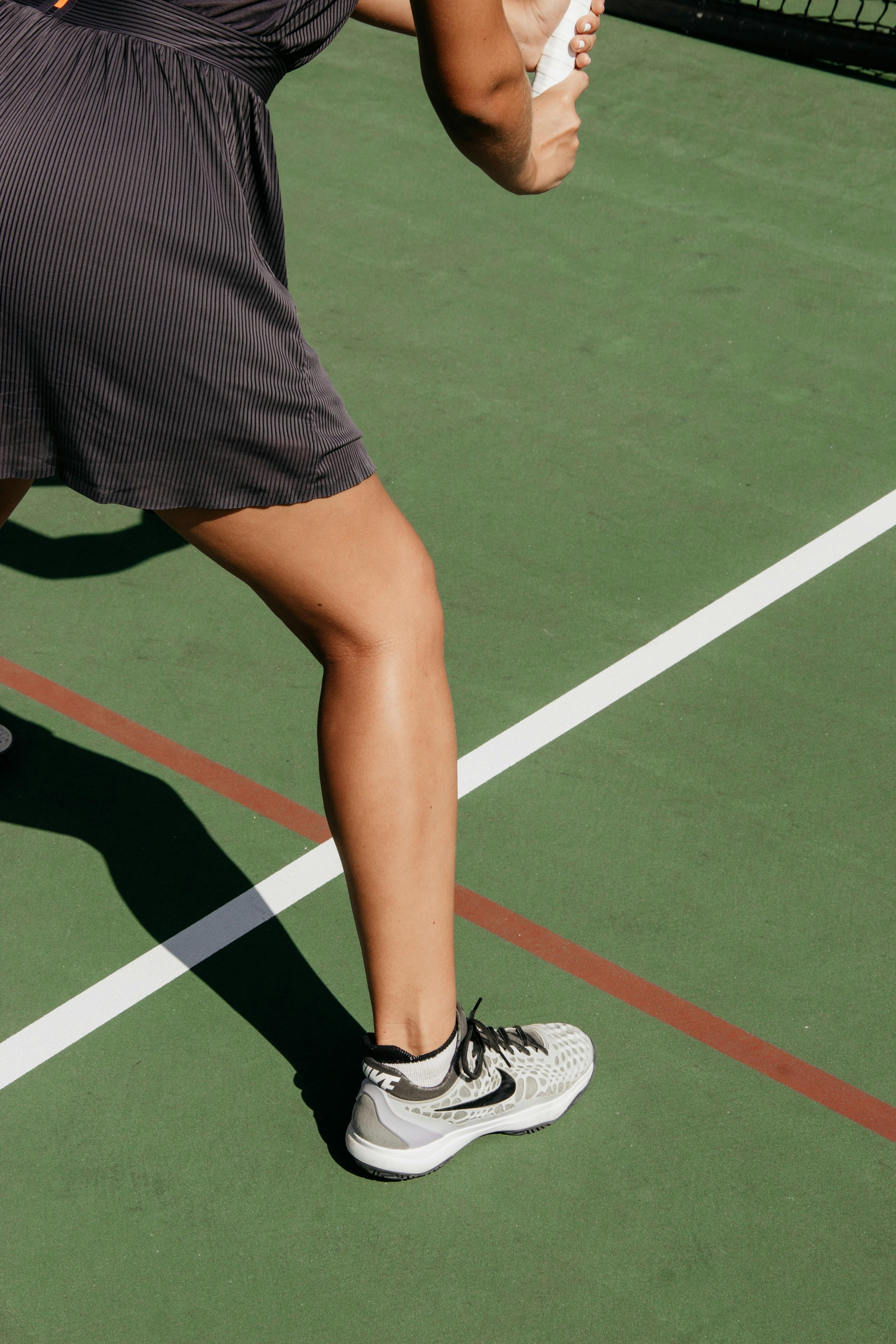 A woman in a tennis dress stands on a hard court in Nike tennis shoes.