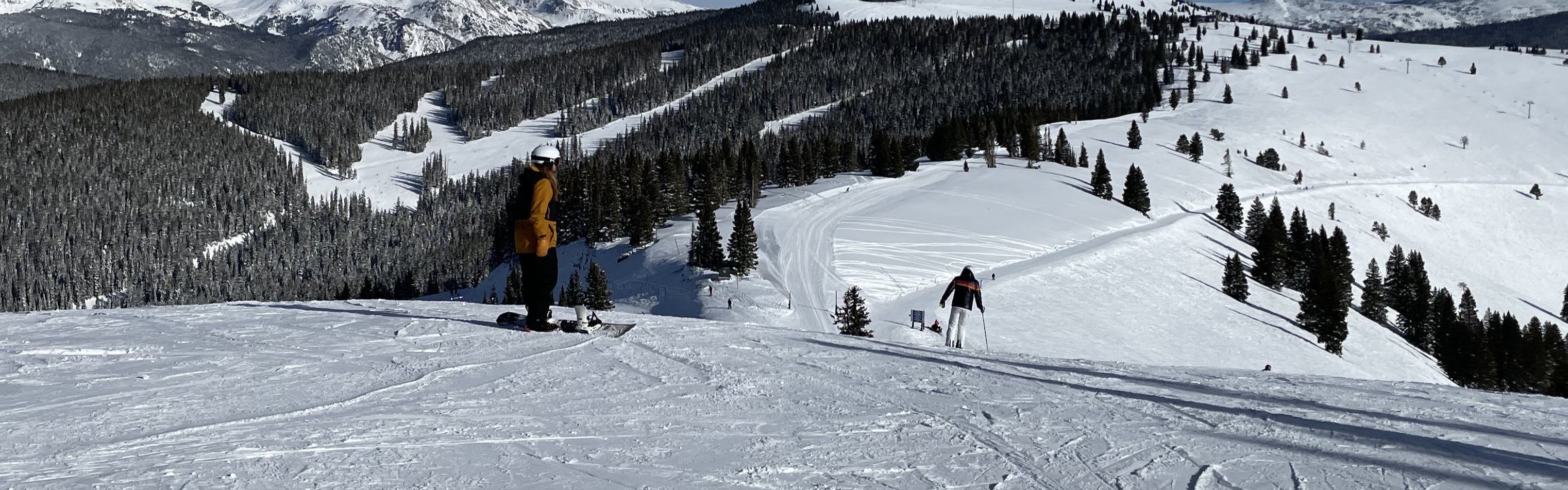 A snowboarder prepared to snowboard down a ski run. There are other skiers and snowboarders visible on the run. 