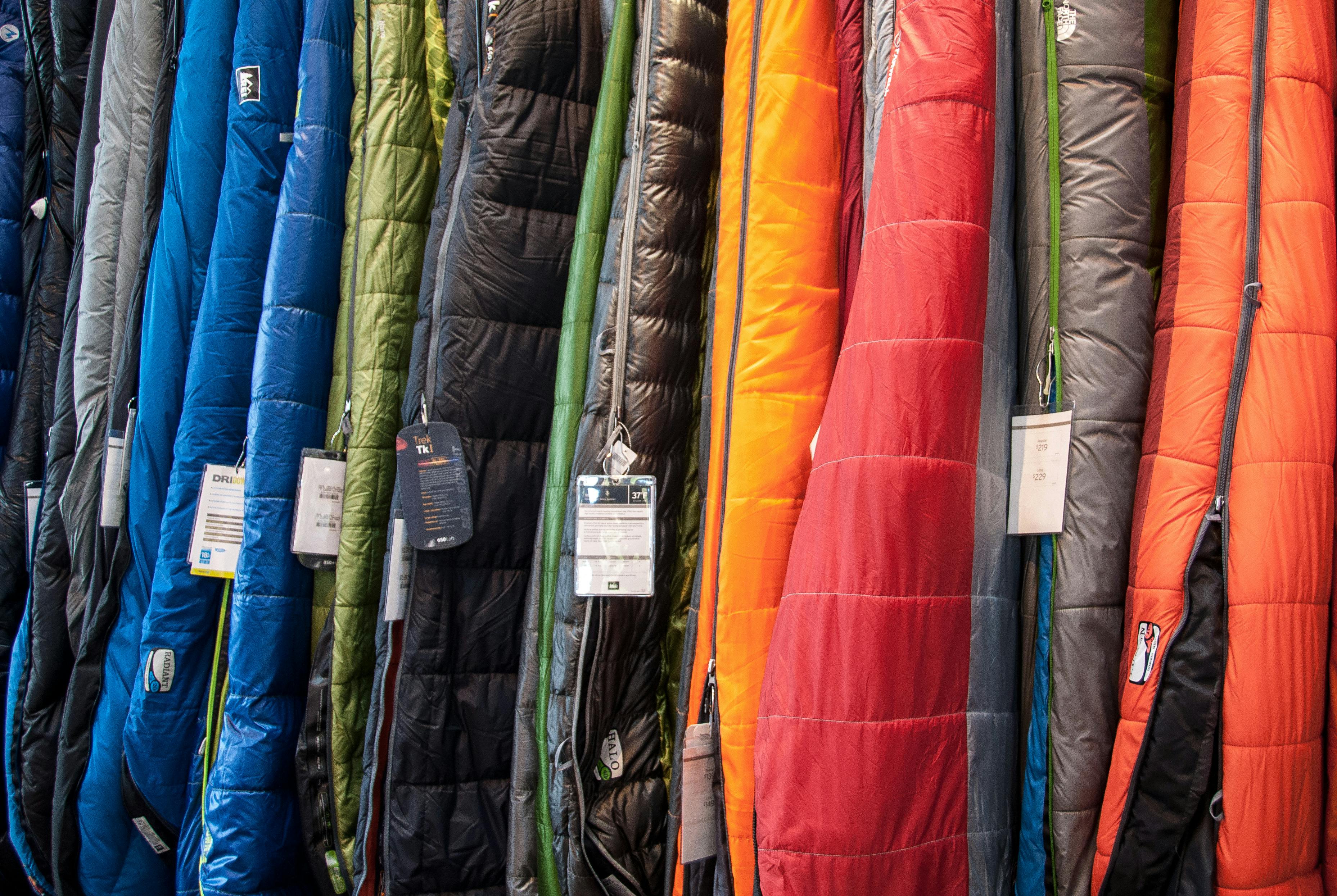 Several sleeping bags in varying colors are hung in a row. They have tags on as if they are still in a store.