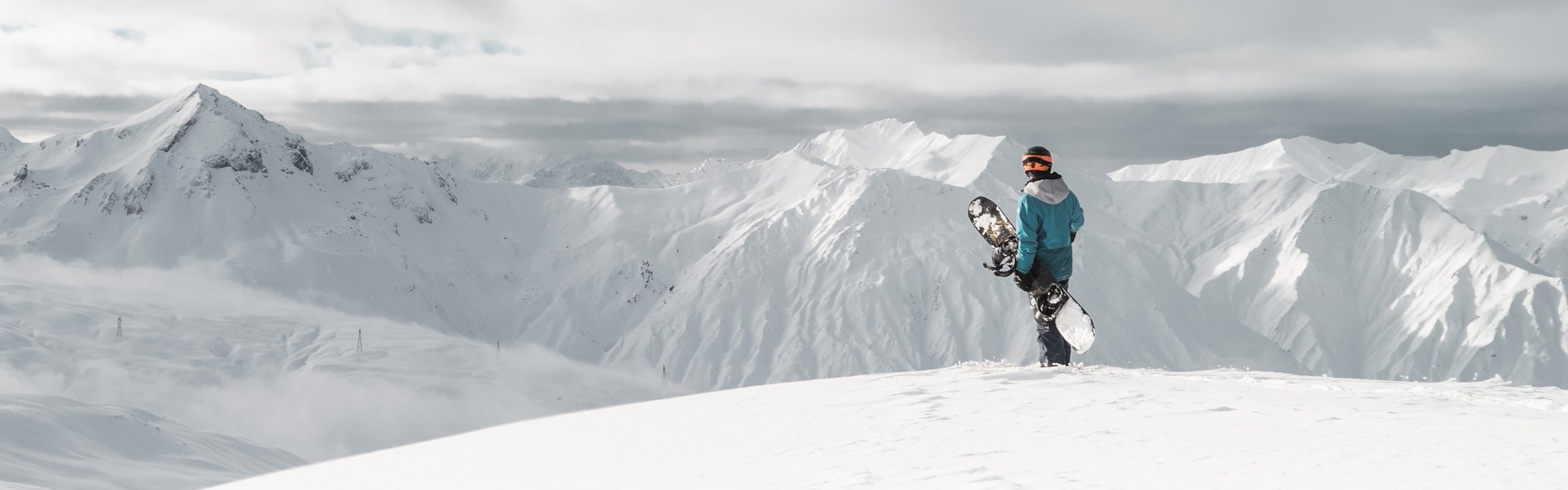 A snowboarder stands at the top of a run and looks down at snowy mountains