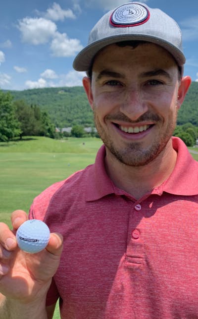 A man holds the Callaway 2021 SuperSoft Golf Ball while smiling. There is a golf course visible behind him.