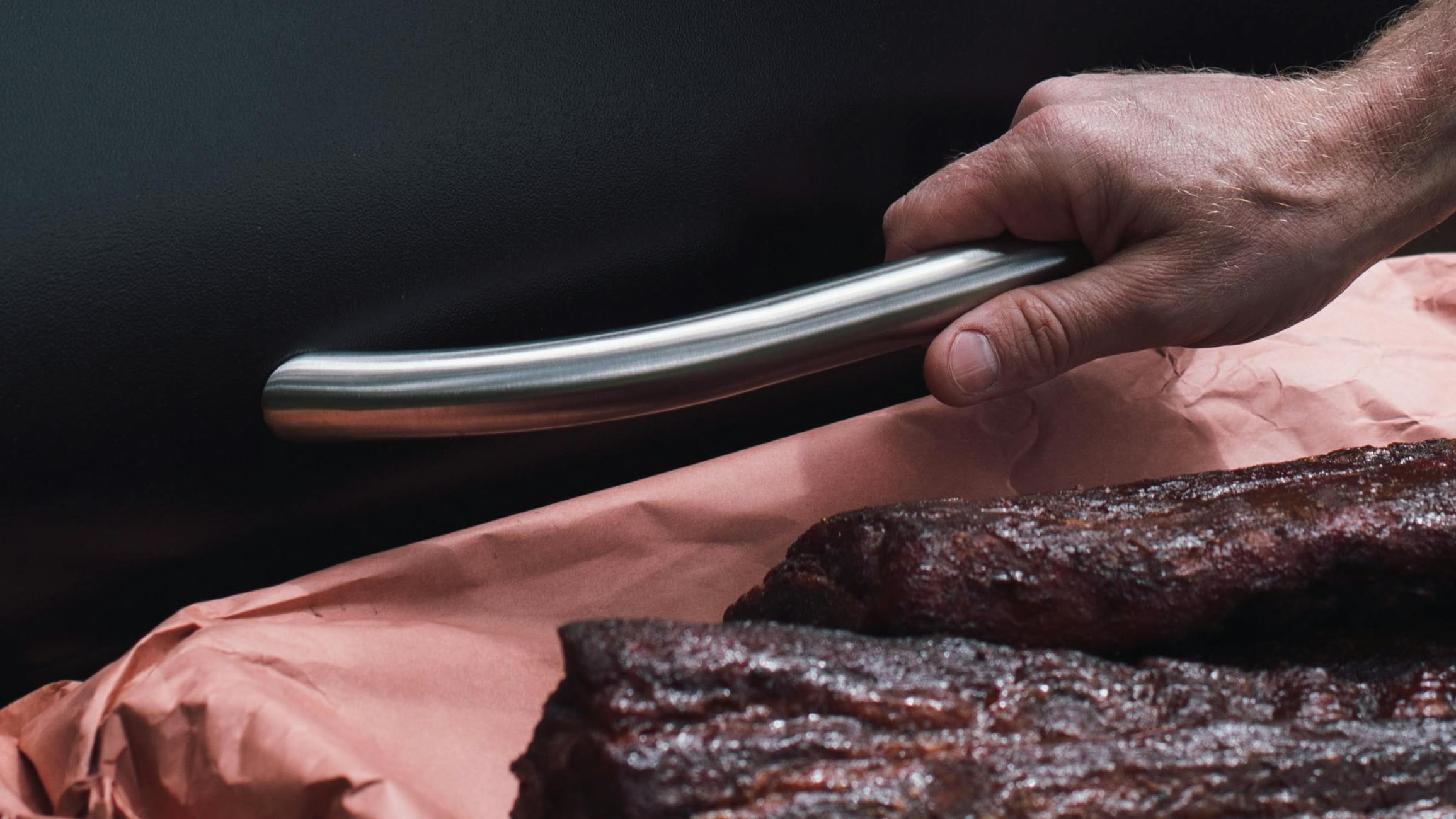 A hand opens a pellet grill with one hand while holding a board of meat with the other hand.
