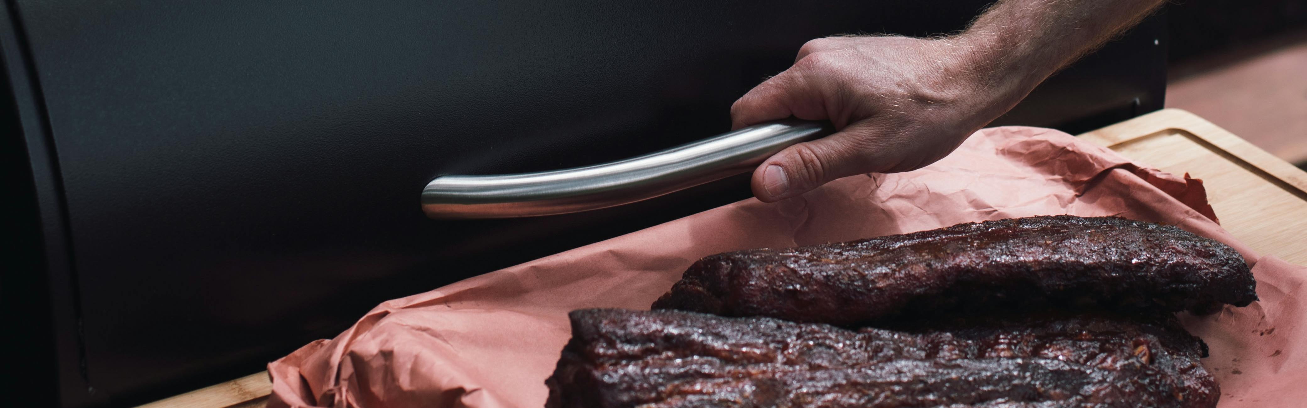 A hand opens a pellet grill with one hand while holding a board of meat with the other hand.