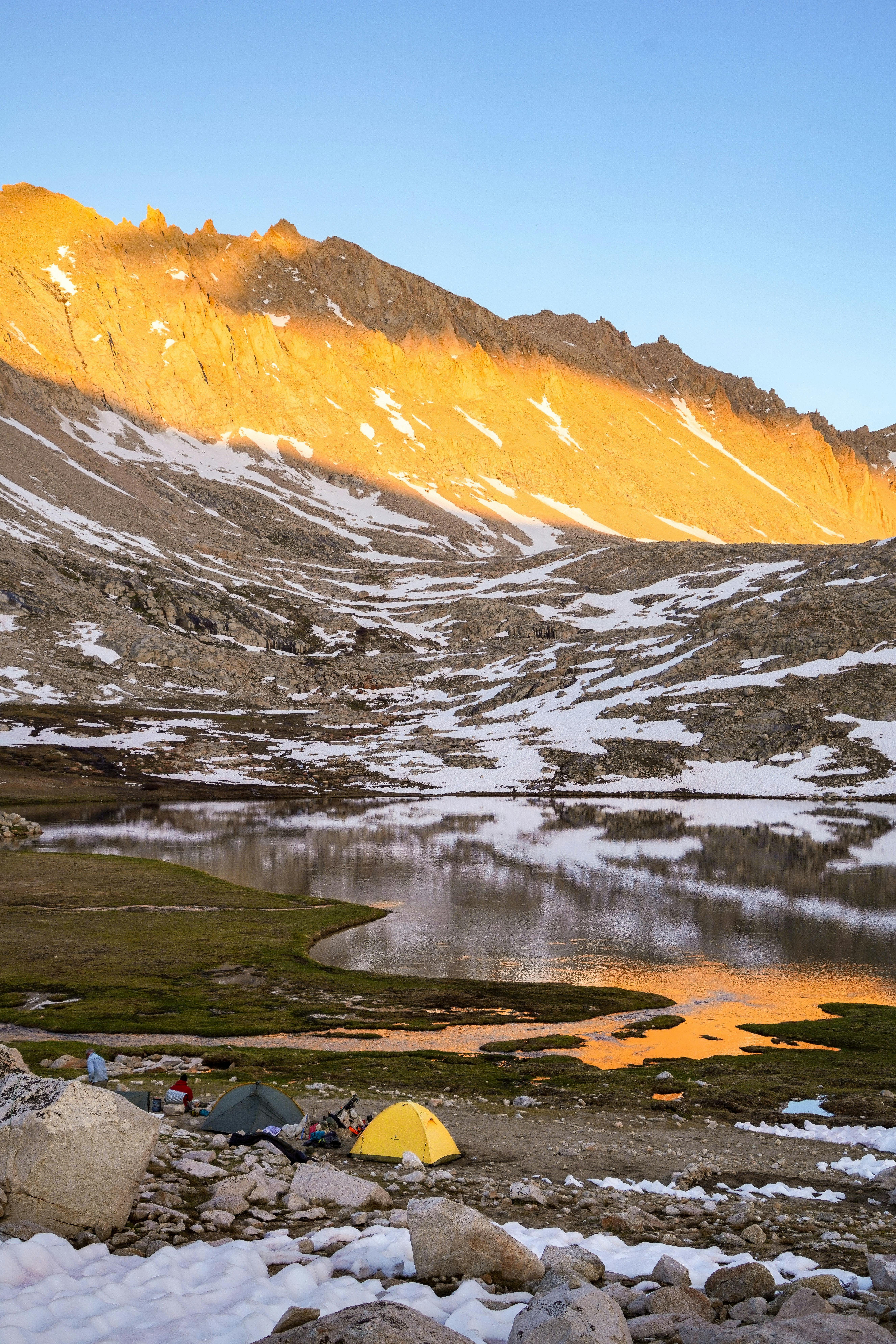 Two small tents are in the bottom of the image, on the shore of a lake that is reflecting the yellow sunset light shining on the jagged, snow-dusted peaks above. 