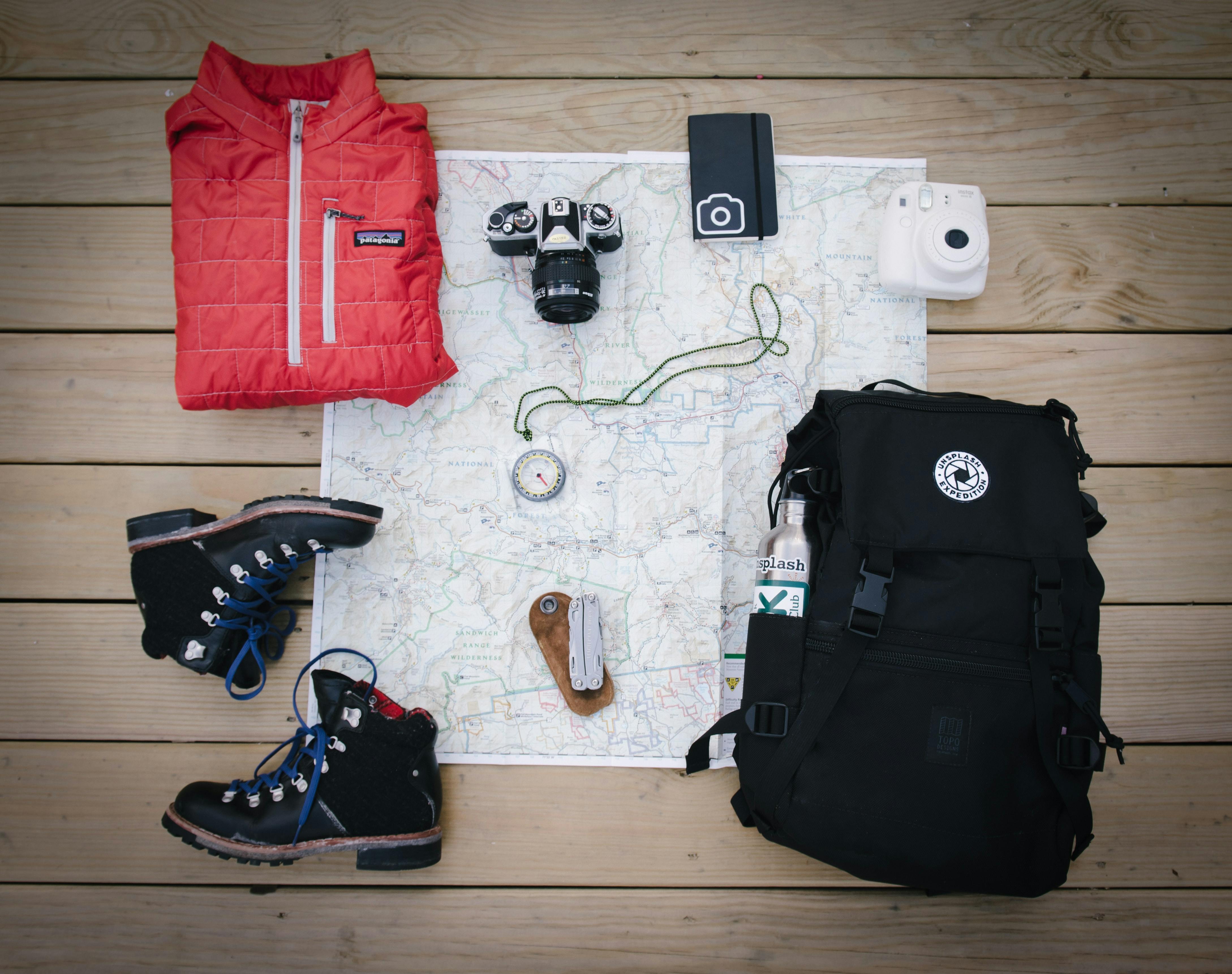 A map, hiking boots, red jacket, black backpack, camera, compass, journal, and camera on the ground