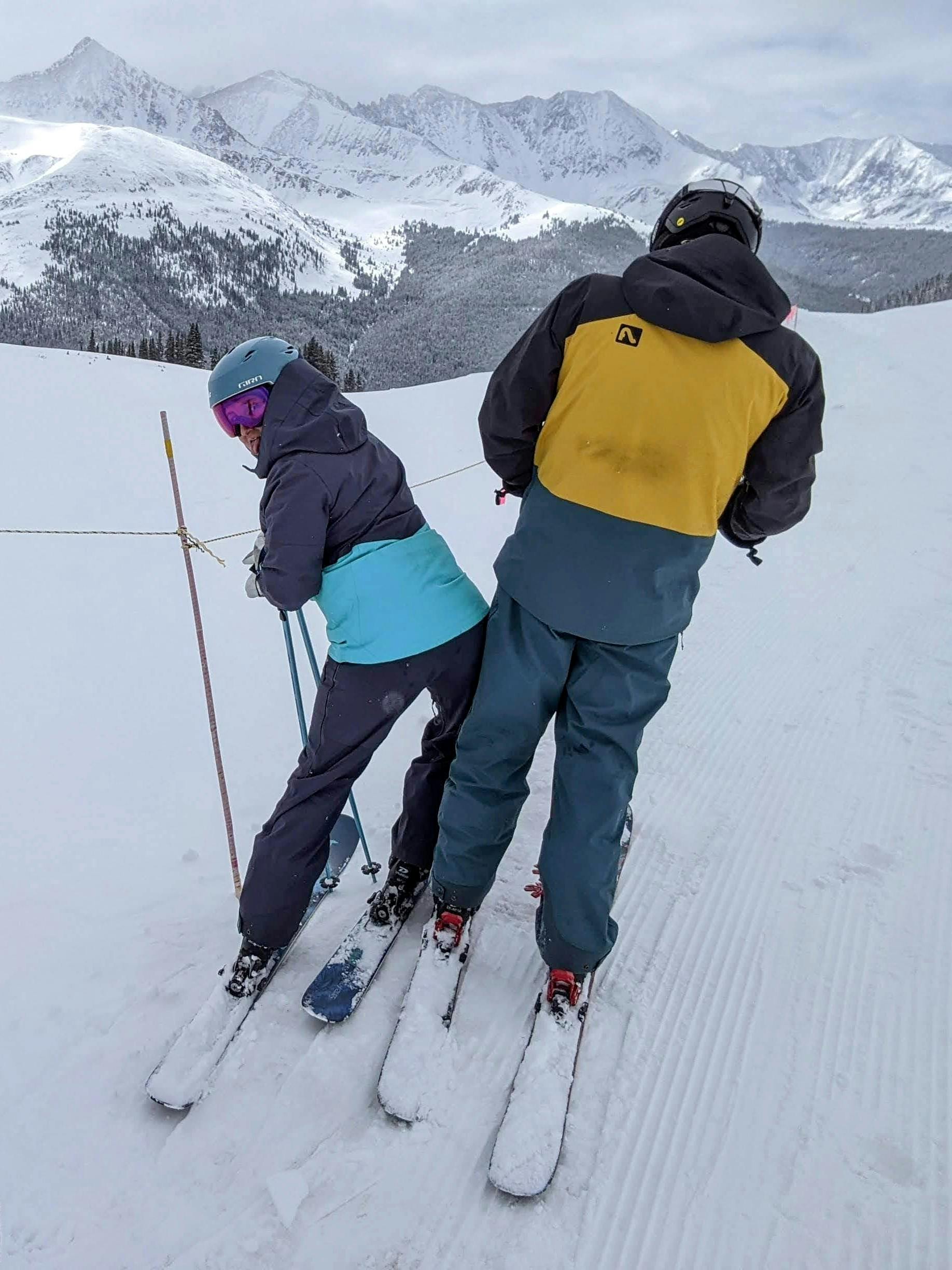 Two skiers standing at the top of a snowy ski run. There are snow capped mountains in the background. 