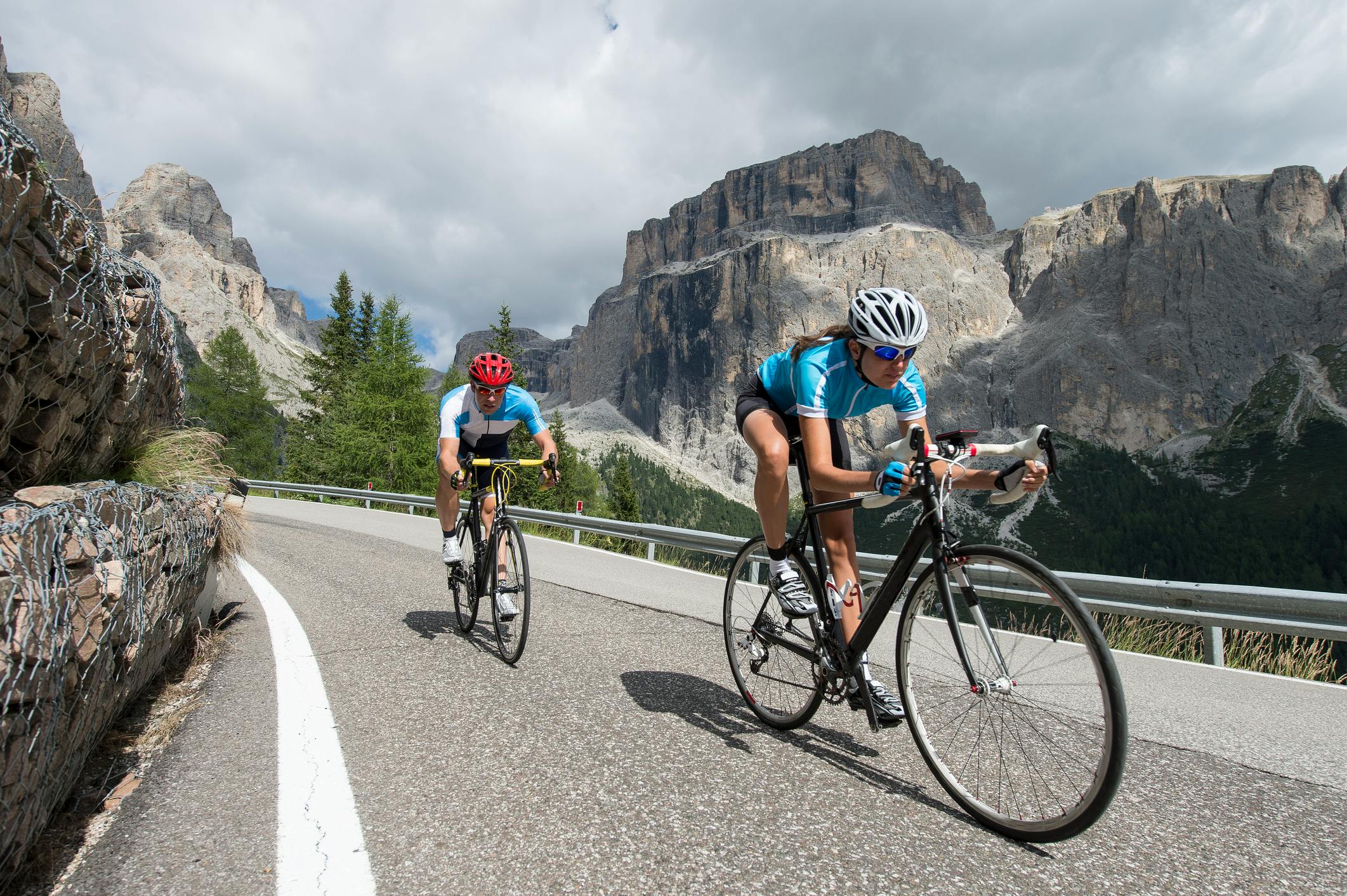 Two cyclists ride up a paved road with mountains in the background