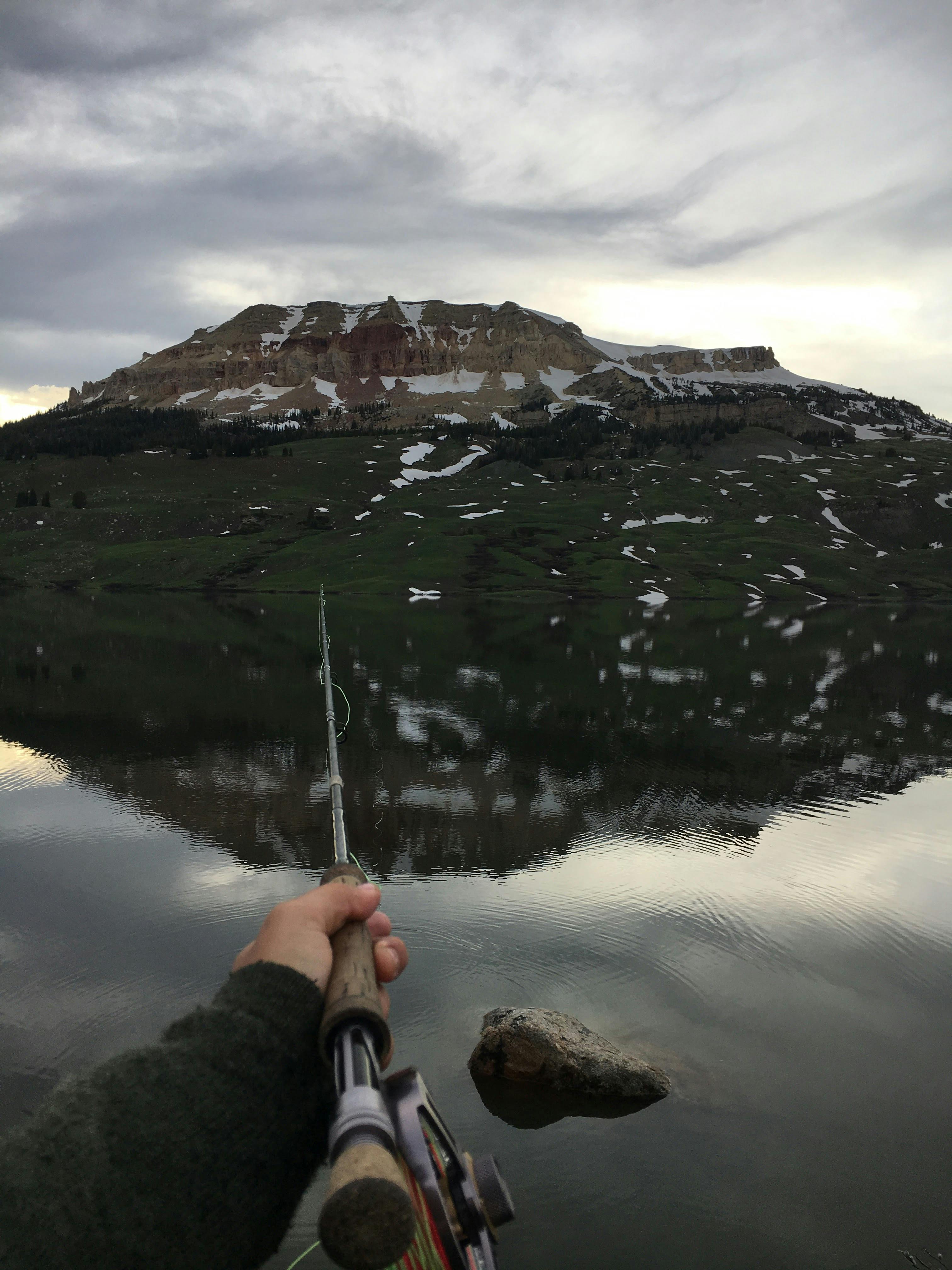 A person casts a fly rod out onto reflective water with a mountain across the way