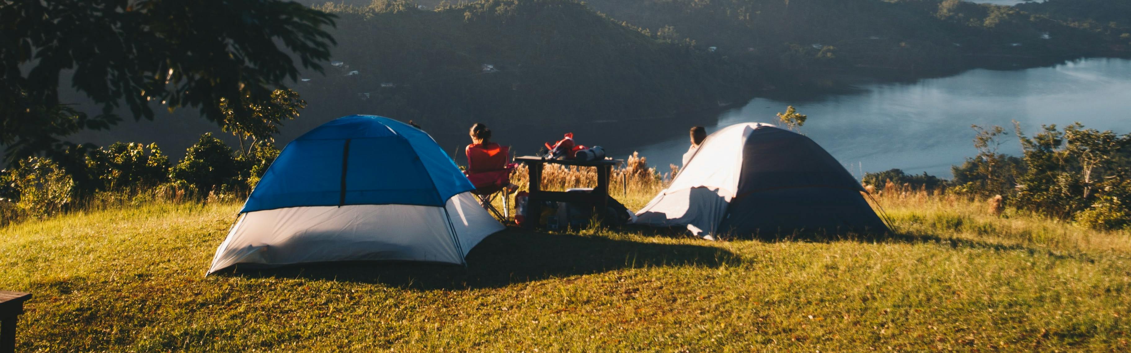 Two tents set up in a grassy field overlooking a body of water. There are two campers sitting outside the tents in camp chairs, and a camp table between the tents. 