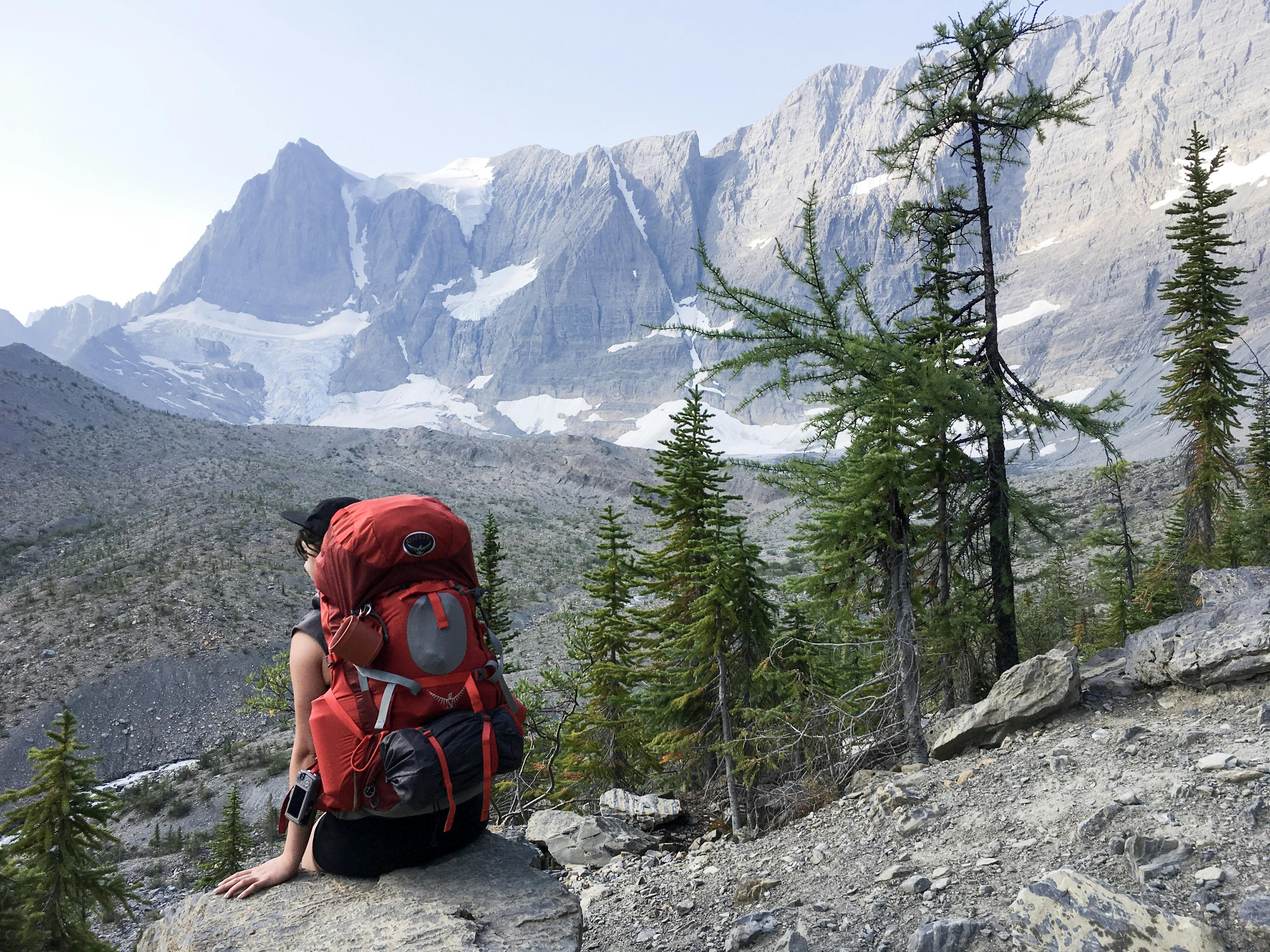 A woman with a red Osprey Ariel 65 Backpack sits on a lookout. There are some rocks around and snowy mountains in the distance.