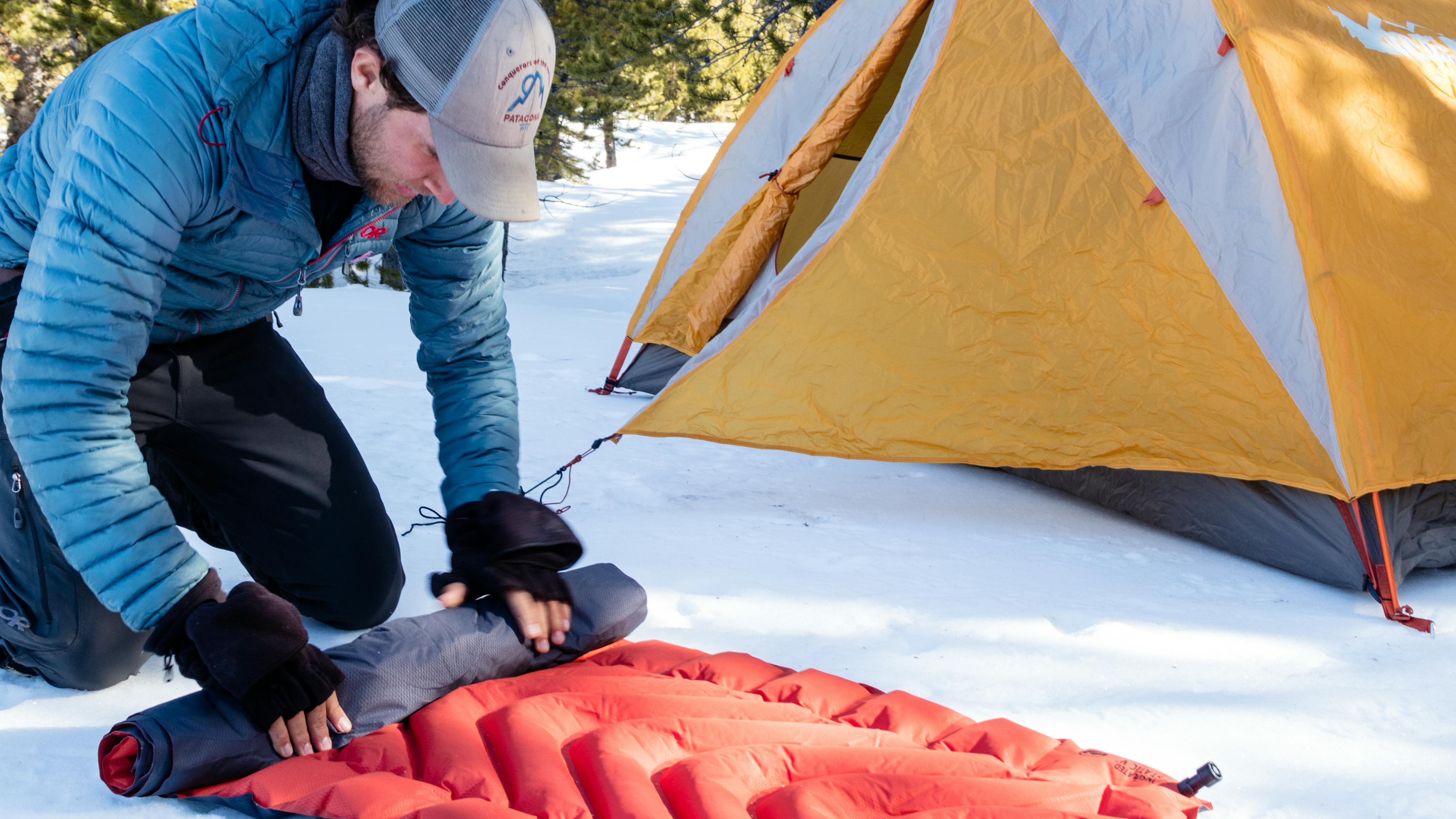A man rolling up a sleeping pad. There is snow on the ground and a tent set up nearby.