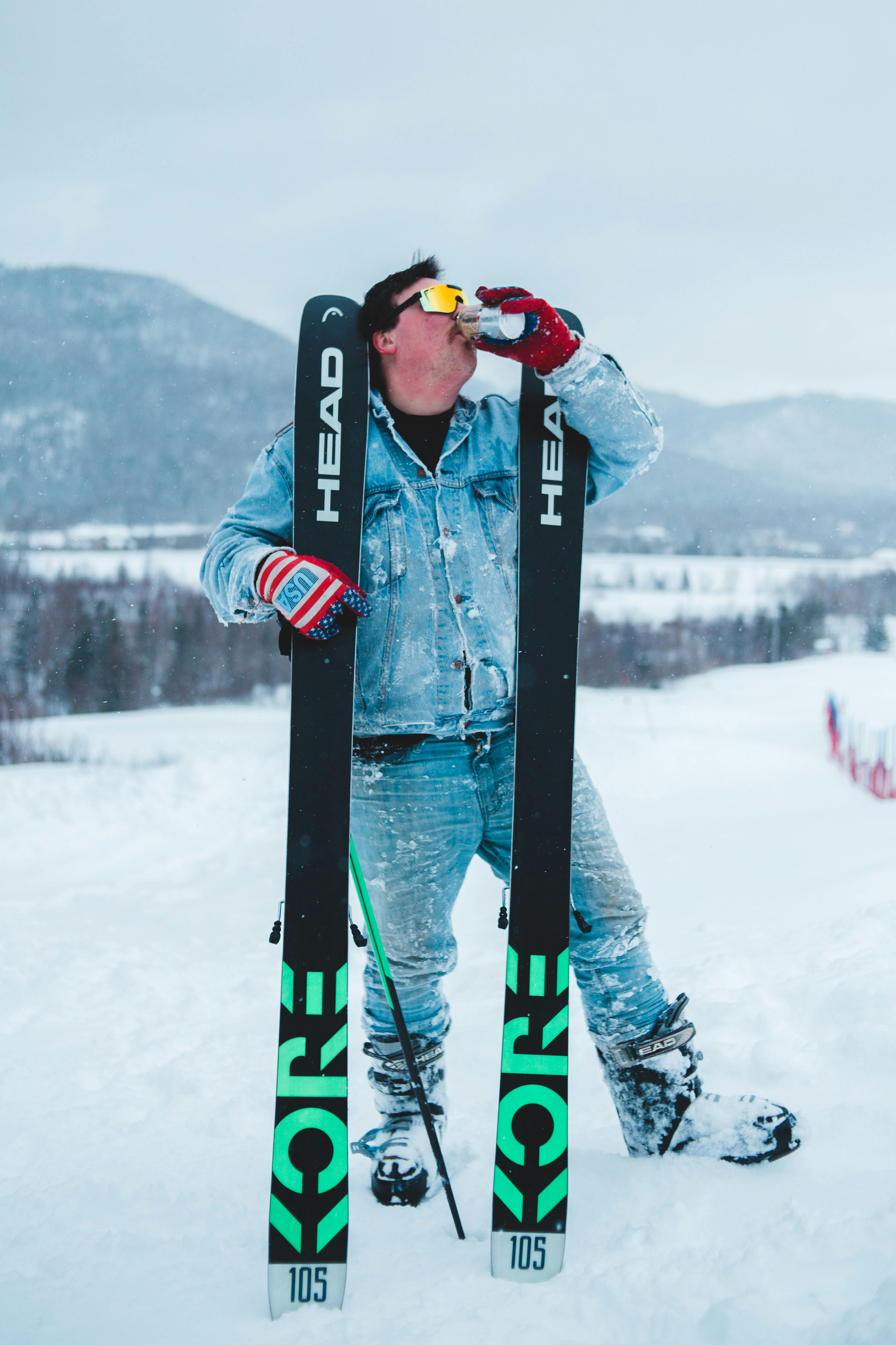 A man wearing jeans and a denim jacket holds up skis while drinking a beer