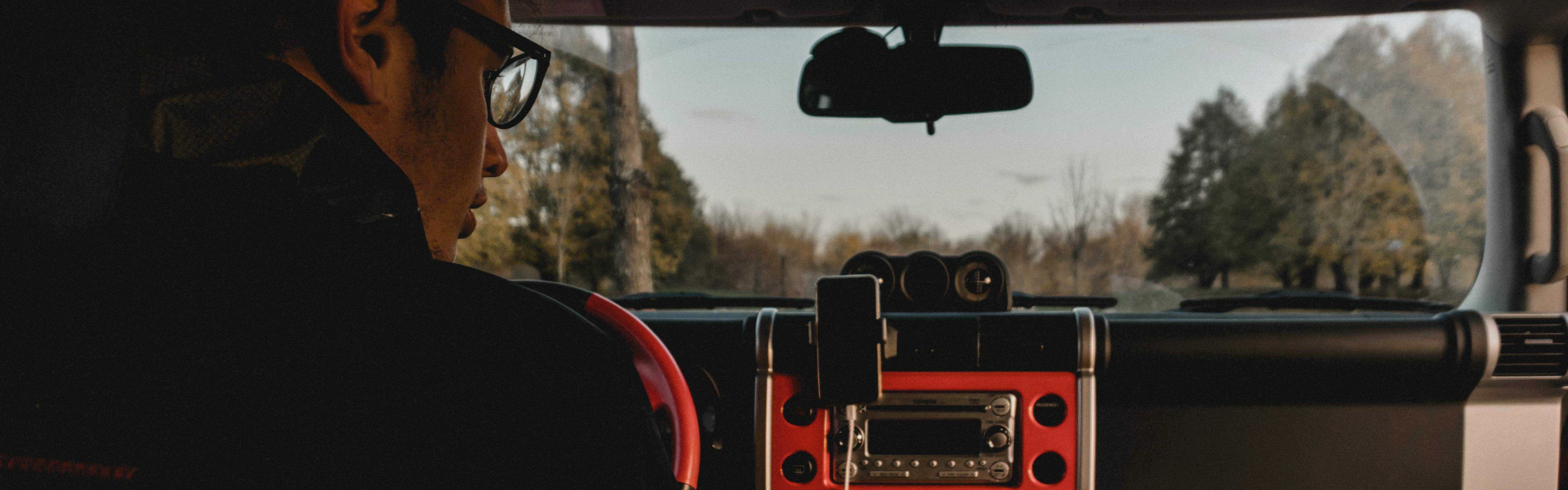 Man listens to something on the car radio as he is driving through a forest area. The radio is red and trees can be seen outside the window.
