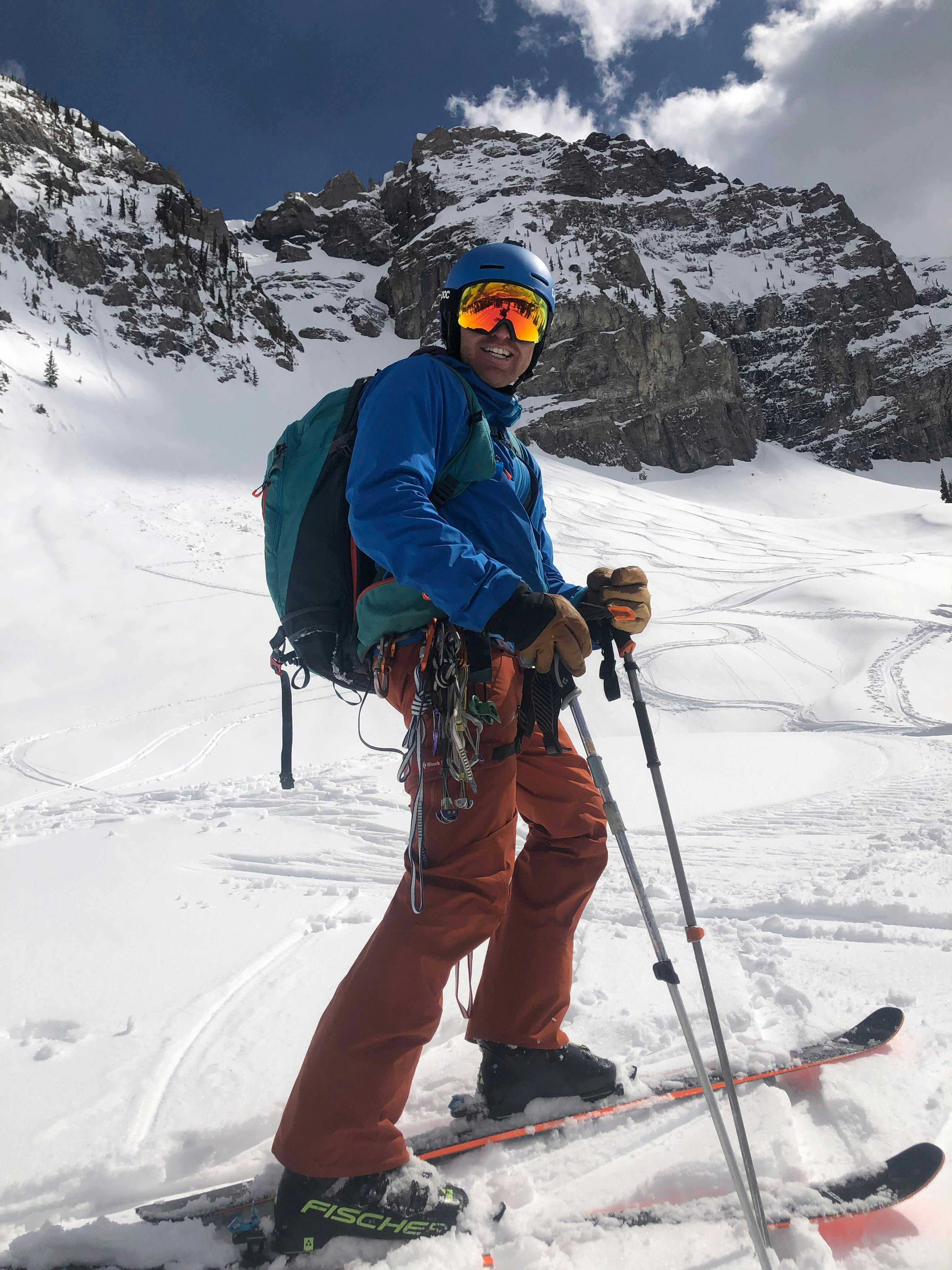 A man in ski gear stands at the bottom of a run. He also has a harness and a bunch of gear on. 