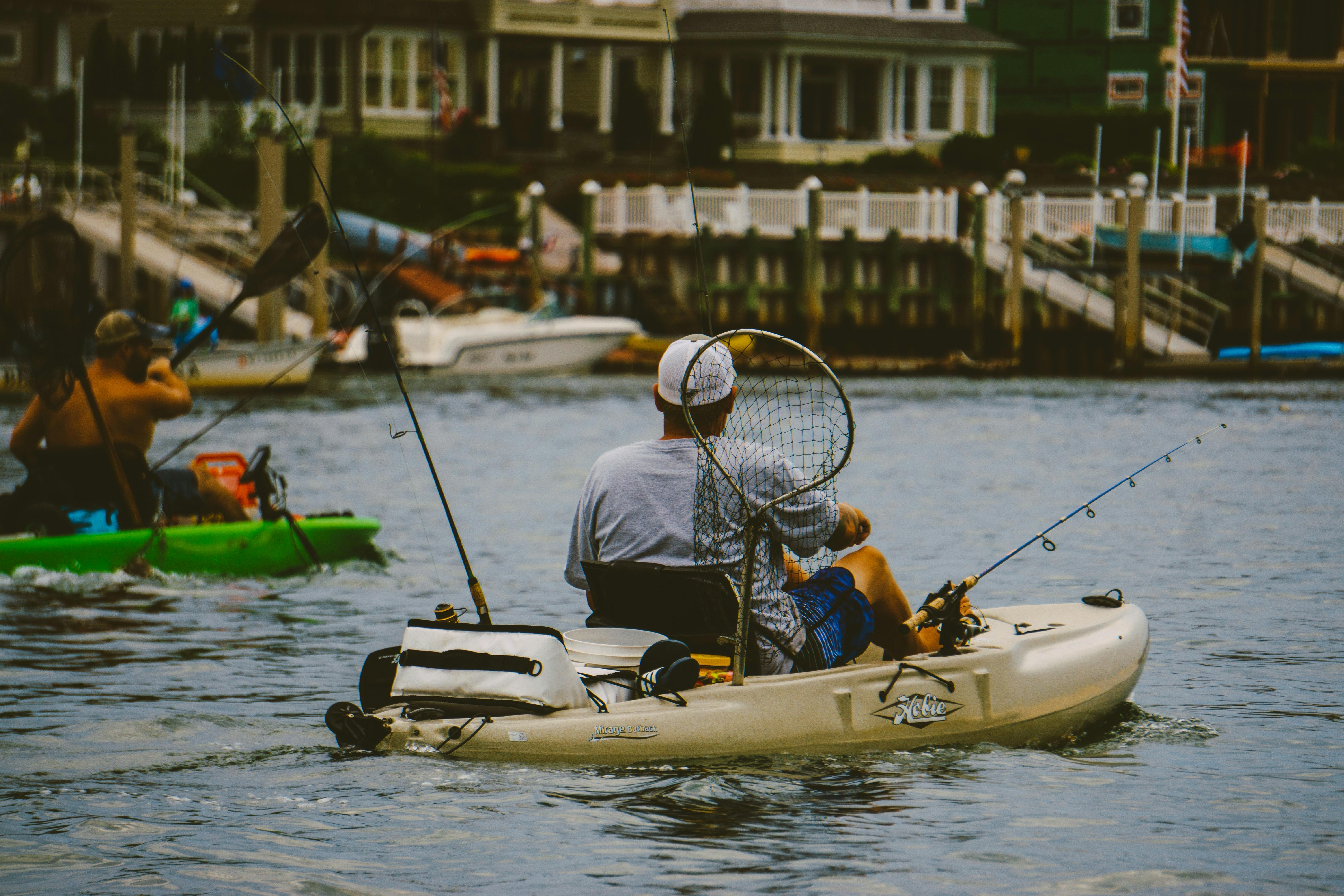 A man kayaks low to the water with a cooler, net, bucket, and fishing rod all visible from the boat.