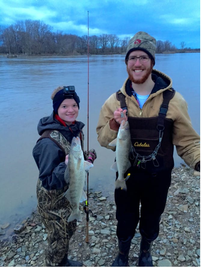 The author wears waders and stands next to a man also in waders. They are both smiling and holding up walleyes that they have caught. 