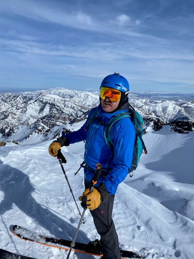 A skier stands wearing all his ski gear. There is a mountain range in the background.