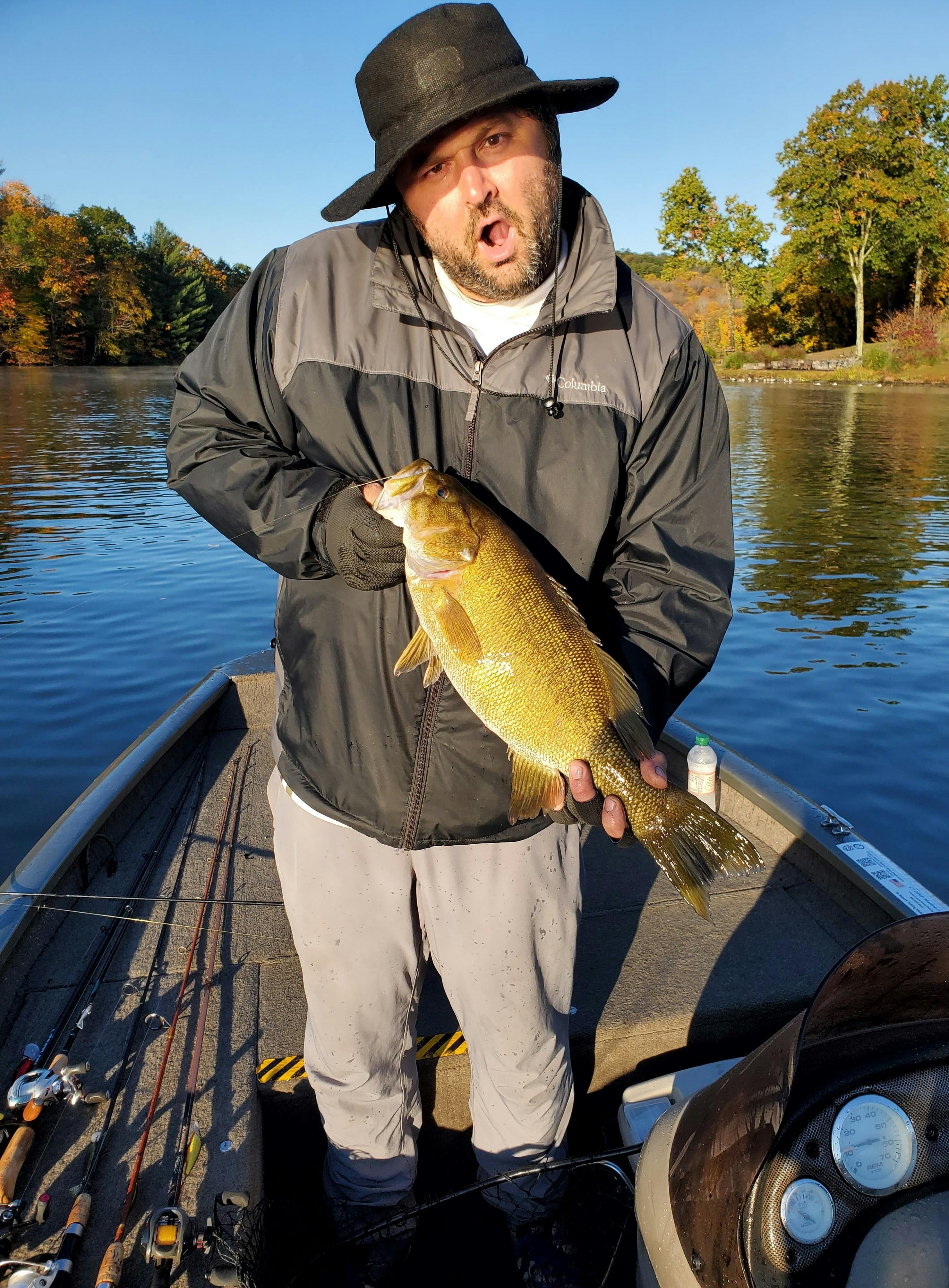 A man holds a smallmouth bass and makes a face, feigning shock. He is standing on a boat in a lake. 