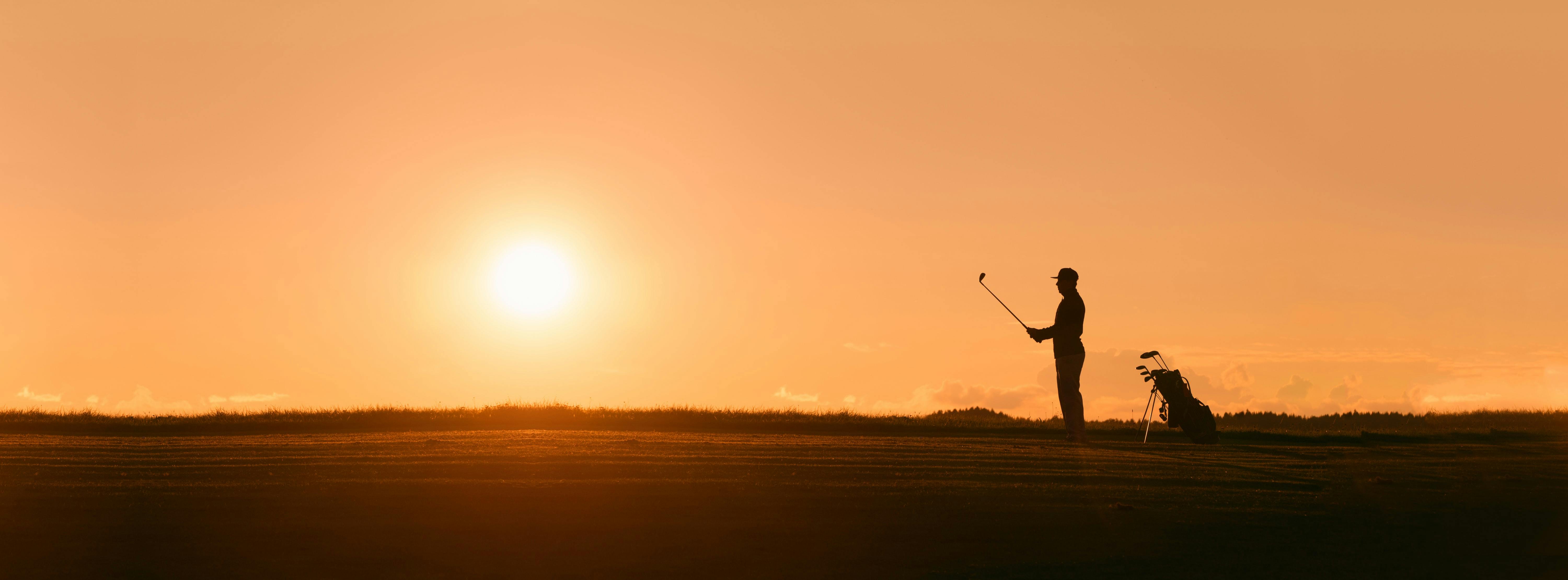 A golfer stands with his club as the sun is going down.