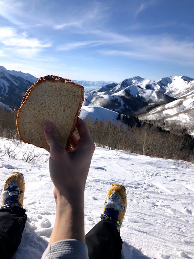 Someone holds up a sandwich and takes a picture of their outstretched legs sitting on the snowy ground. 
