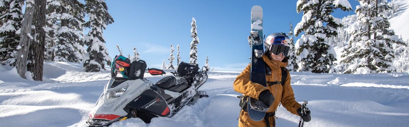 Tatum Monod carries her skis and walks away from a snowmobile in deep powder. 
