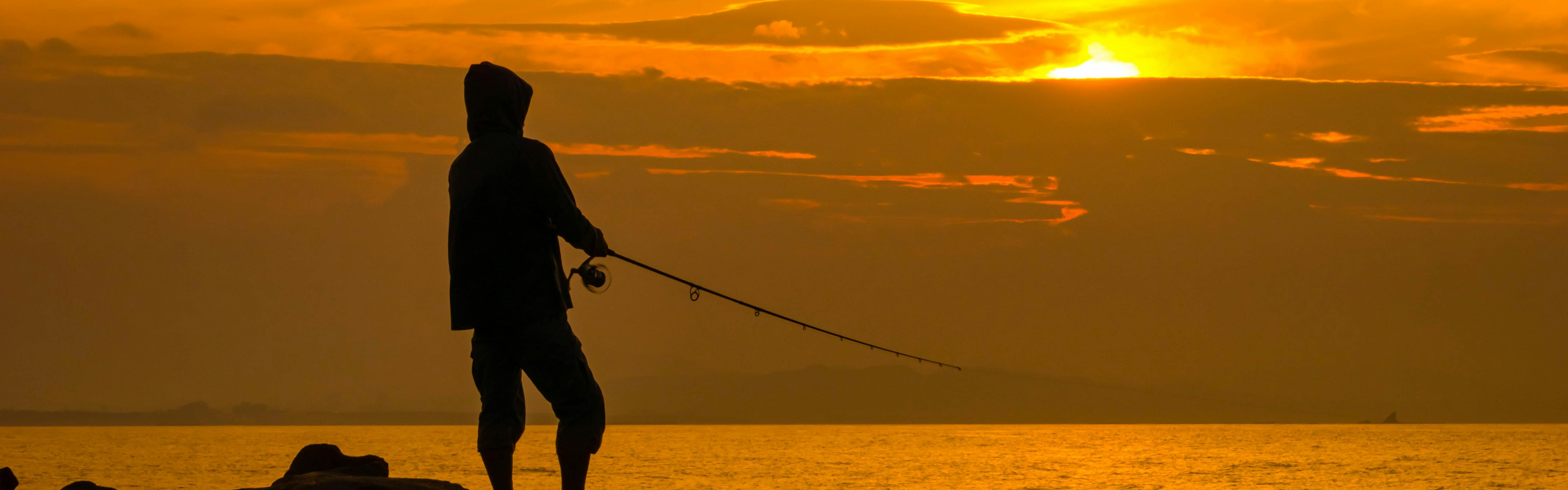 A silhouette of a man fishing backdropped by a glowing, orange sunrise. 