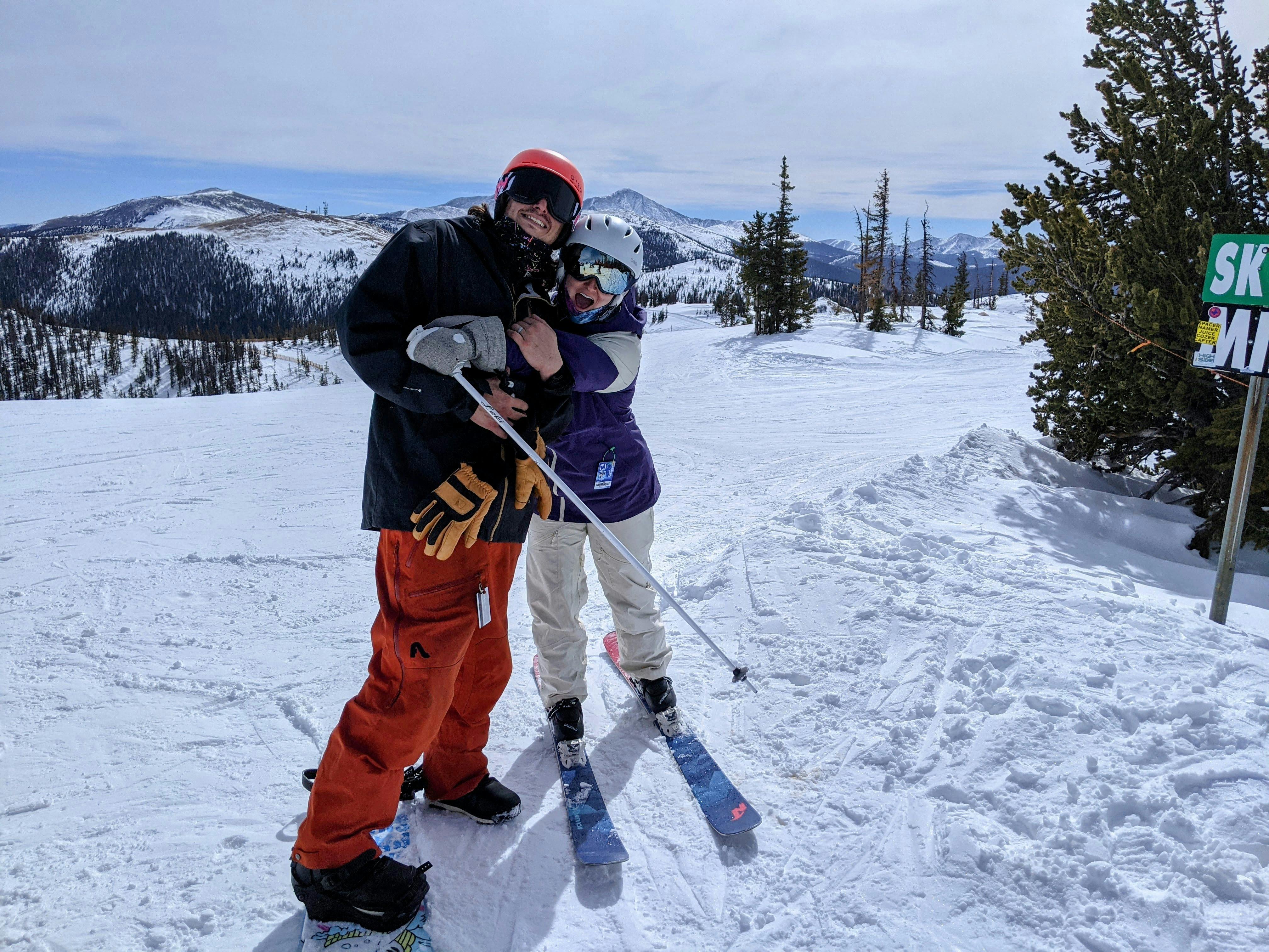 Two people on a snowy ski trail, one is wearing skis and hugging the other. 