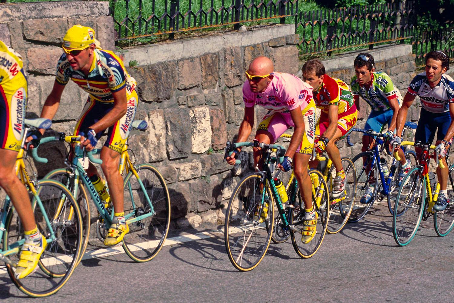 Some road cyclists are racing up a hill. They all have yellow shoes on and some of them are standing up while pedaling. 