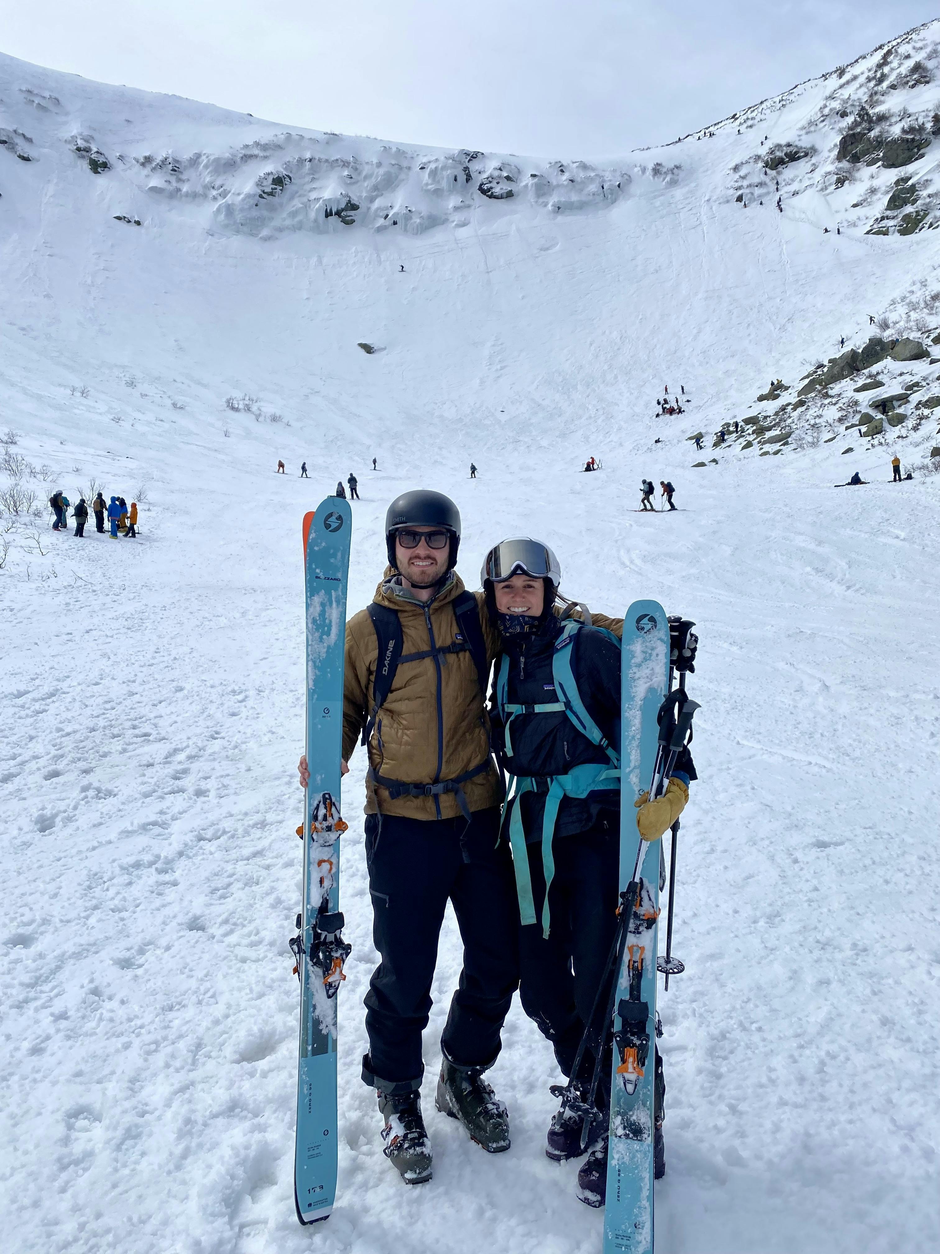 A man and a woman standing at the bottom of a ski area holding their skis next to them. 