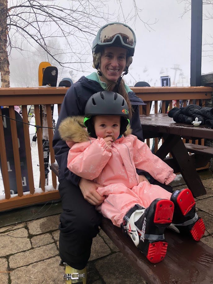 A woman in ski gear holding a child in ski gear. They are sitting at a table outside. 