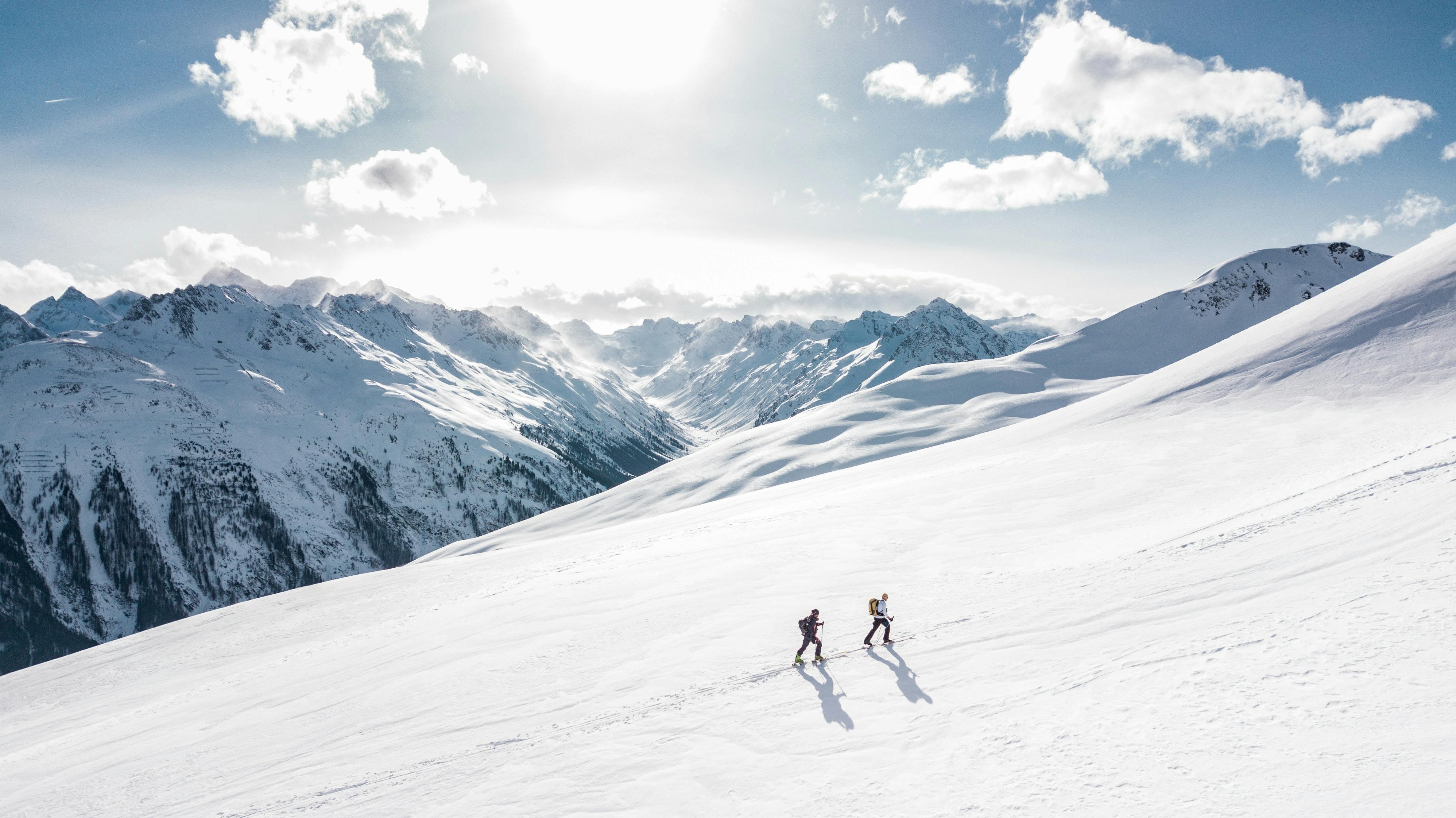 Two skiers walk uphill. The photo is taken from far away and there are some peaks in the background. The sun is shining and snow is everywhere.