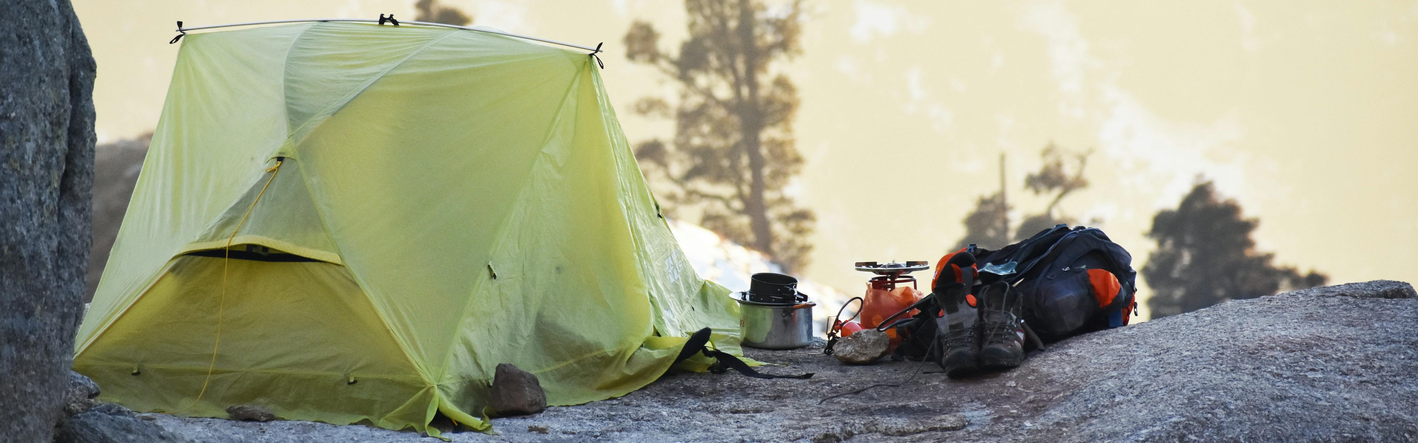 A tent is set up with cooking equipment next to it on a rock base. Snow is visible in the background.