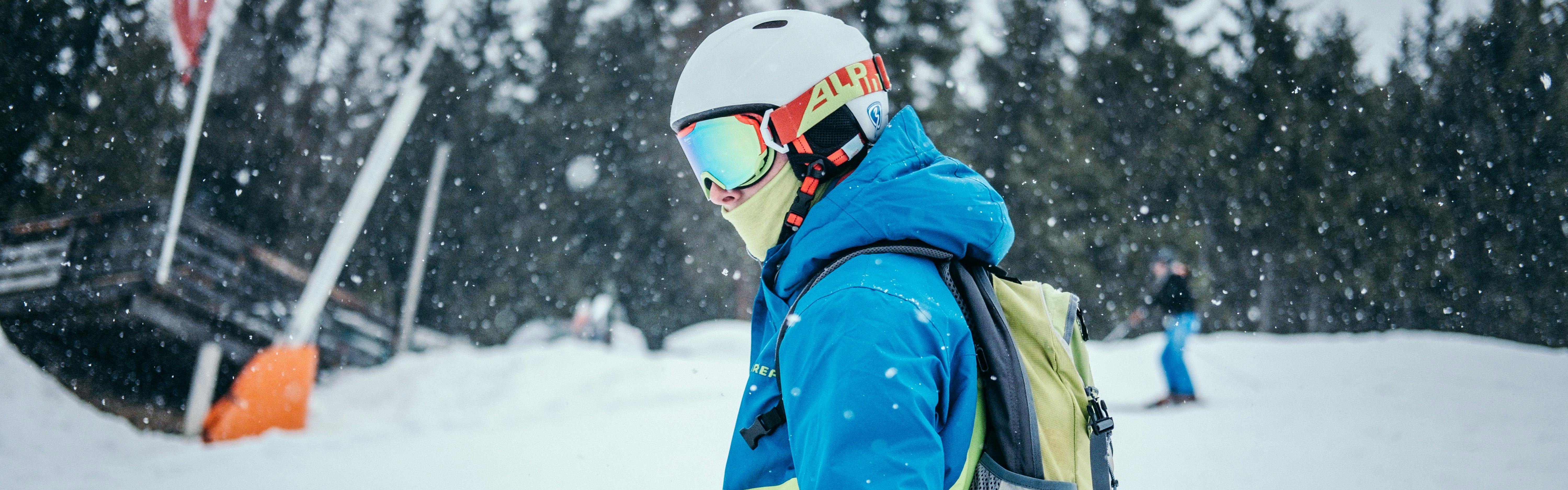 A skier or snowboarder stands wearing a helmet, goggles, and backpack as the snow falls around him. 
