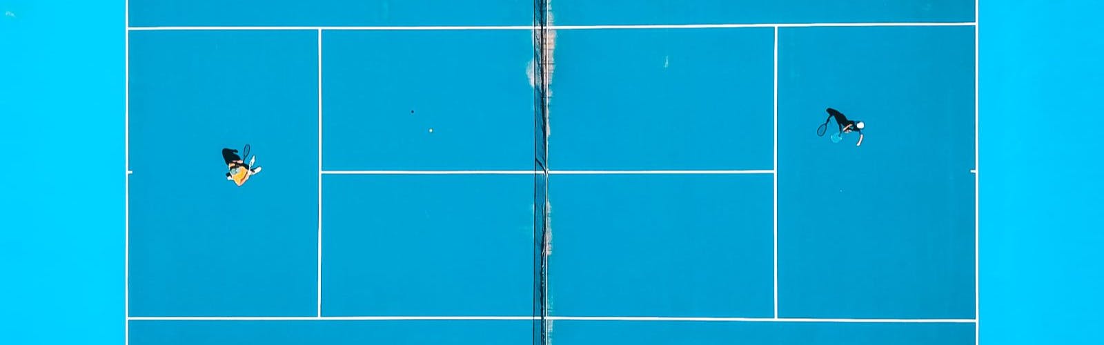 A teal tennis court from above with one person playing on each side of the net.