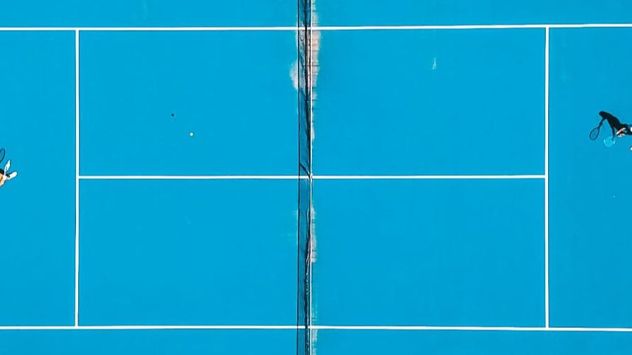 A teal tennis court from above with one person playing on each side of the net.